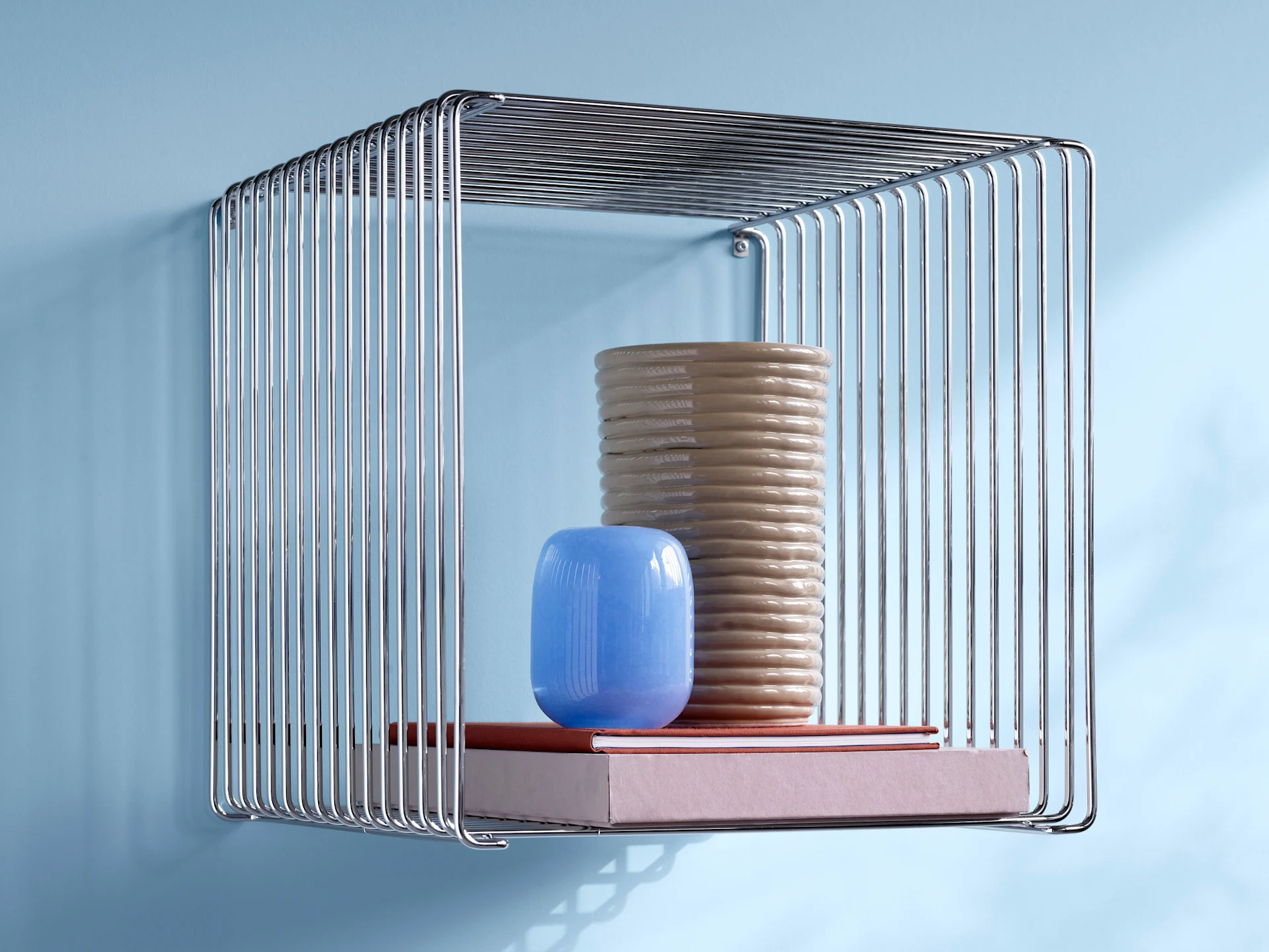 A chrome wire shelf holding a tall, ribbed beige vase, a small blue vase, and two books on a light blue wall.