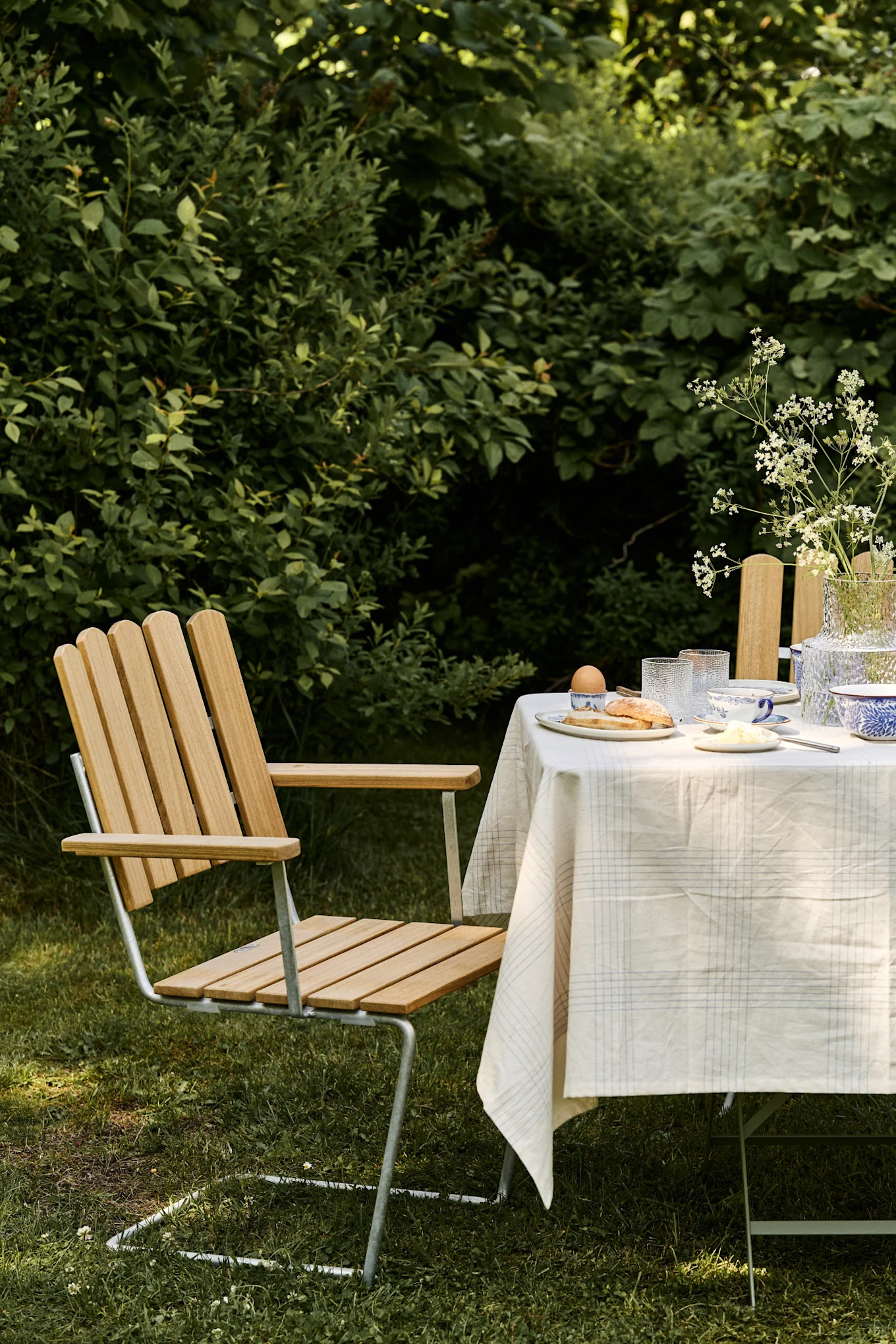 Outdoor dining table with white tablecloth, breakfast items, and wooden slat chair on a green lawn.