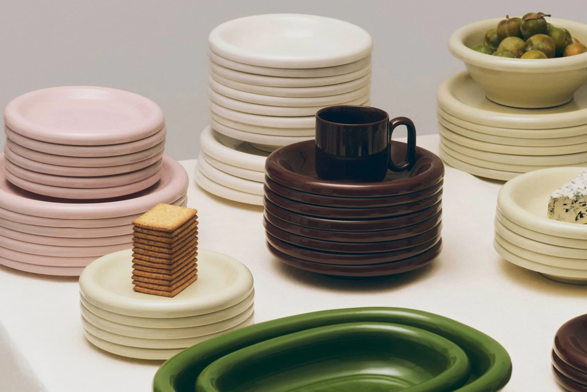 Stacks of round, minimalist plates and bowls in pink, cream, brown, and green, some with olives, cheese, or crackers.
