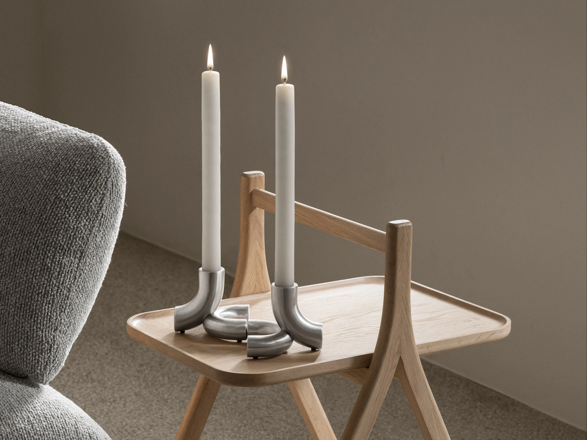 Two lit white candles in curved silver holders on a light wooden side table, with a grey armchair to the left.