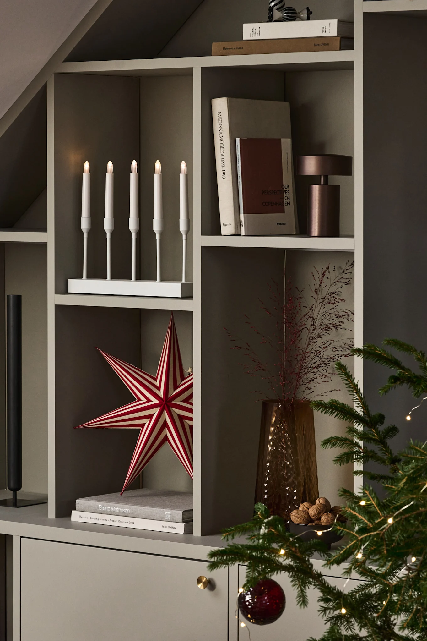A bookshelf filled with Christmas decorations, with a white Advent candle holder glowing on top.