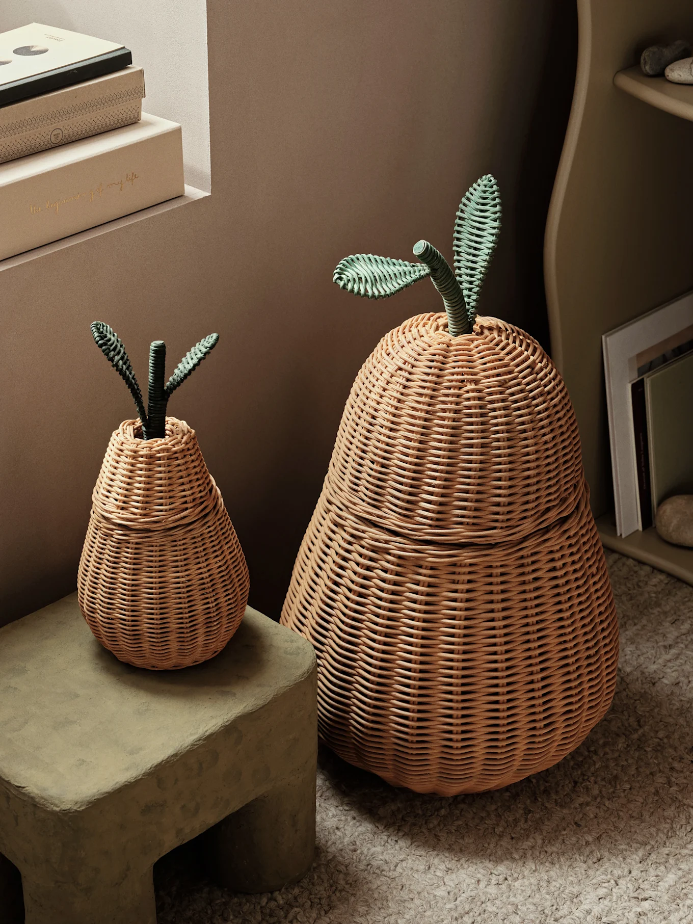 Two light brown woven pear-shaped storage baskets, one large and one small, both with green woven stems and leaves, on a textured stool and rug.