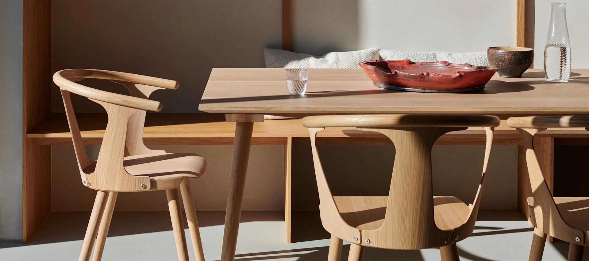 Modern dining area with light wood table and chairs, a red decorative bowl, water glass, and pitcher, bathed in warm sunlight.