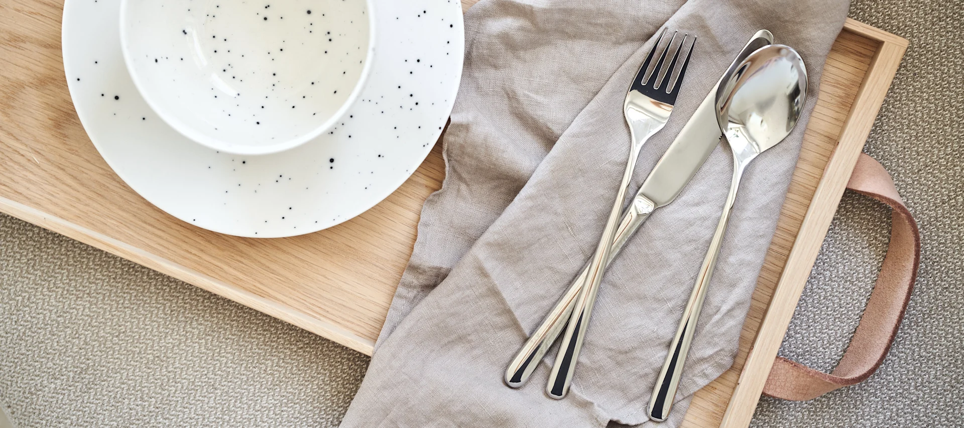 Table setting with a speckled white plate and bowl on a wooden tray, alongside a fork, knife, and spoon on a beige linen napkin.