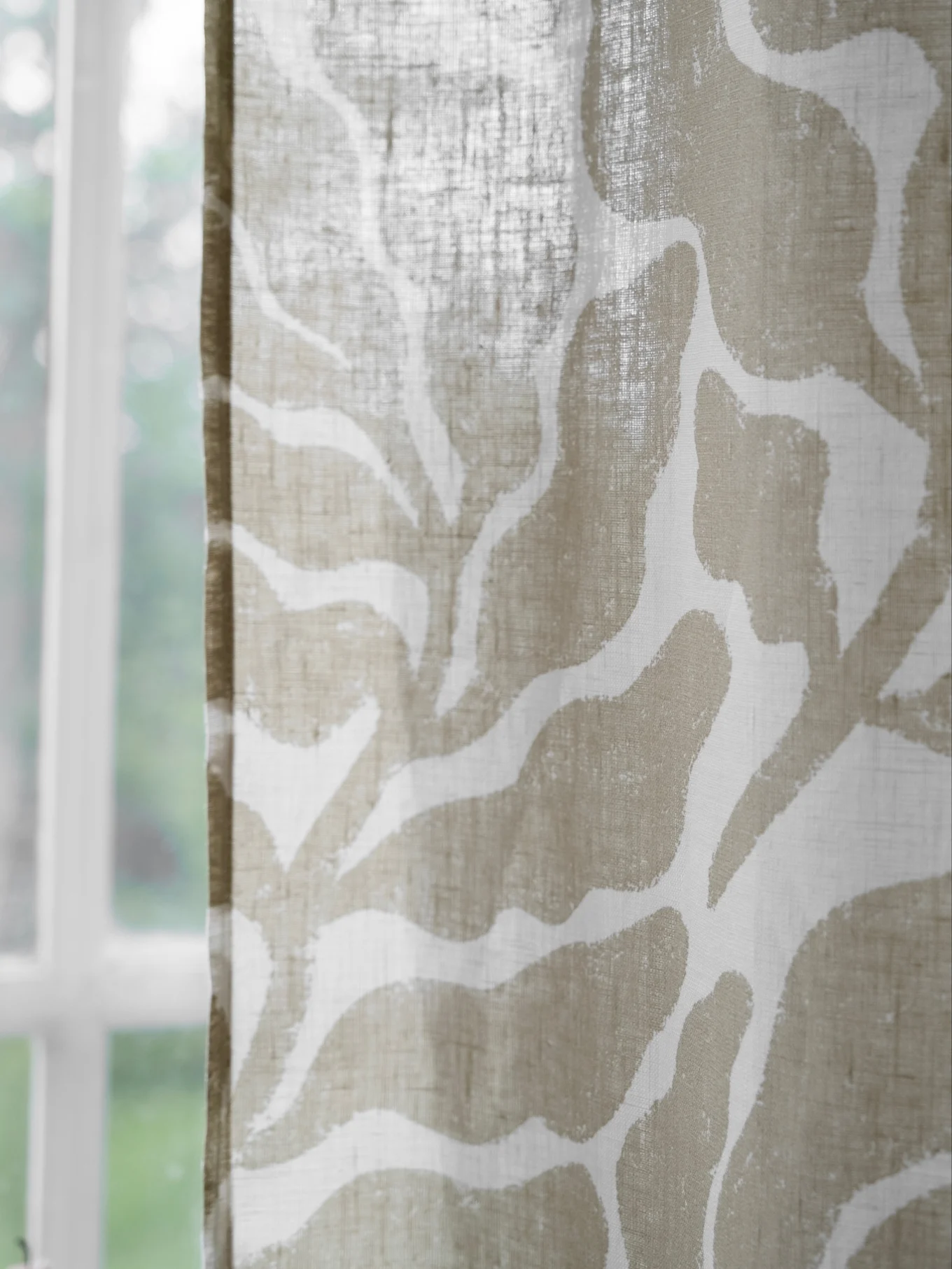 Close-up of a sheer window curtain with a beige abstract pattern, with a blurred window and greenery in the background.