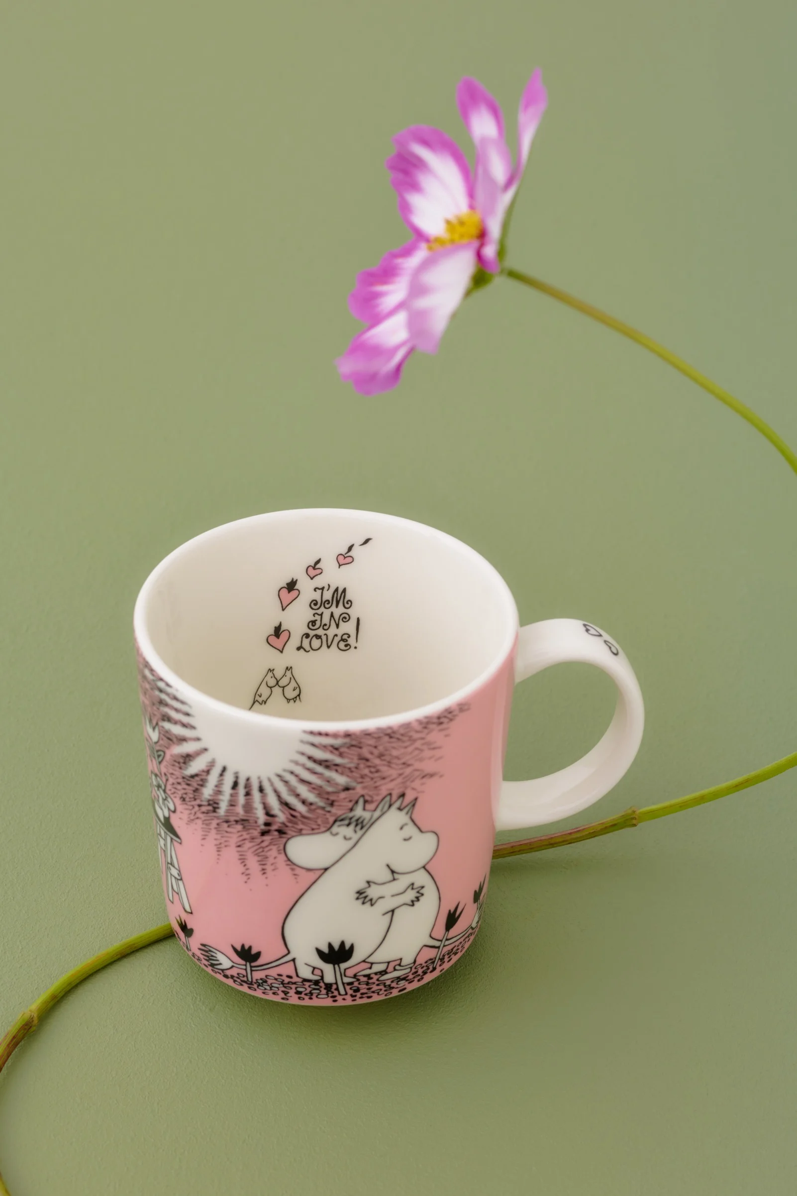 Pink and white Moomin mug with Moomintroll and Snorkmaiden embracing, with "I'M IN LOVE!" text inside, on a green background with a cosmos flower.