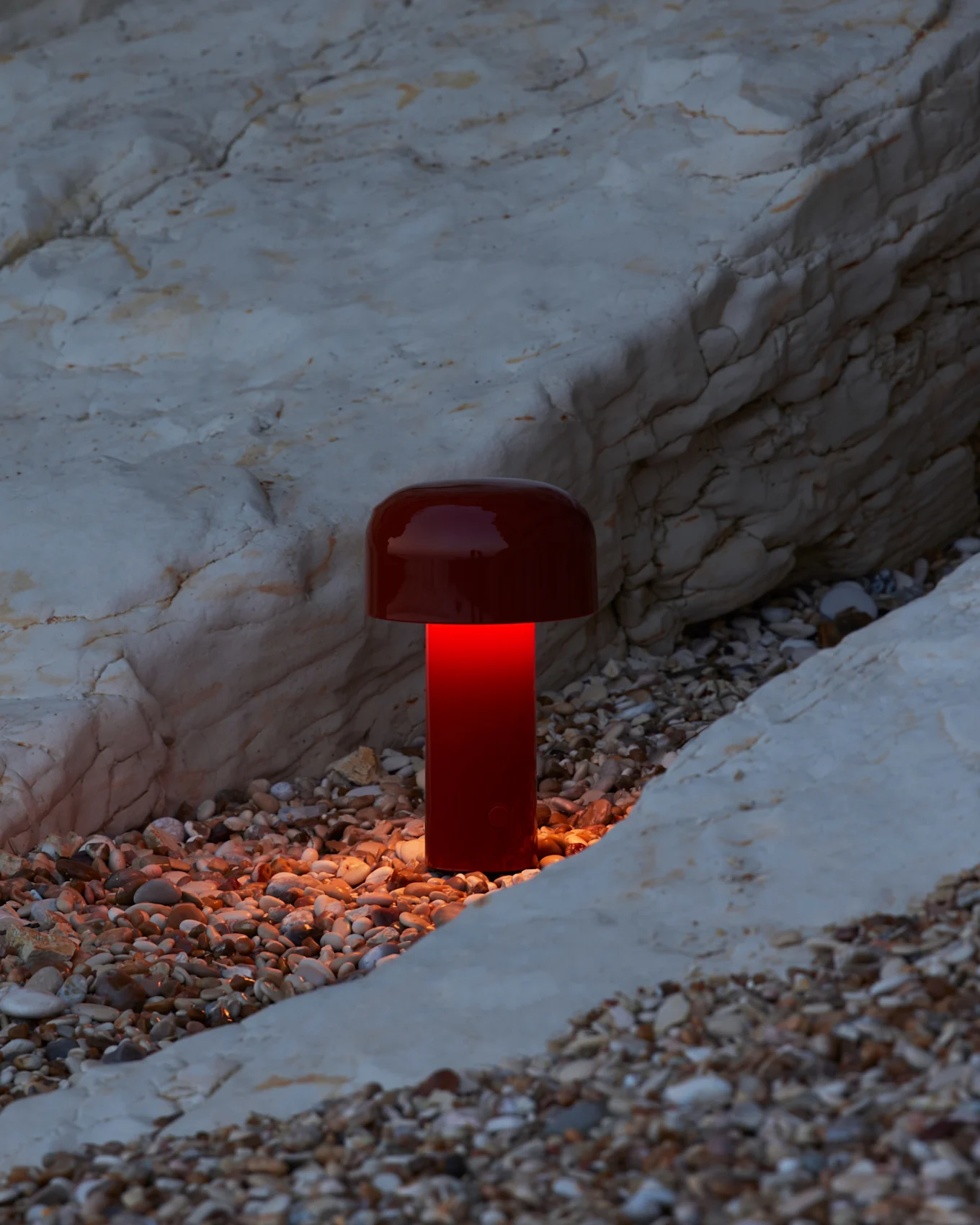 A red mushroom-shaped lamp illuminates a pebble beach with a warm orange glow against pale rocks.