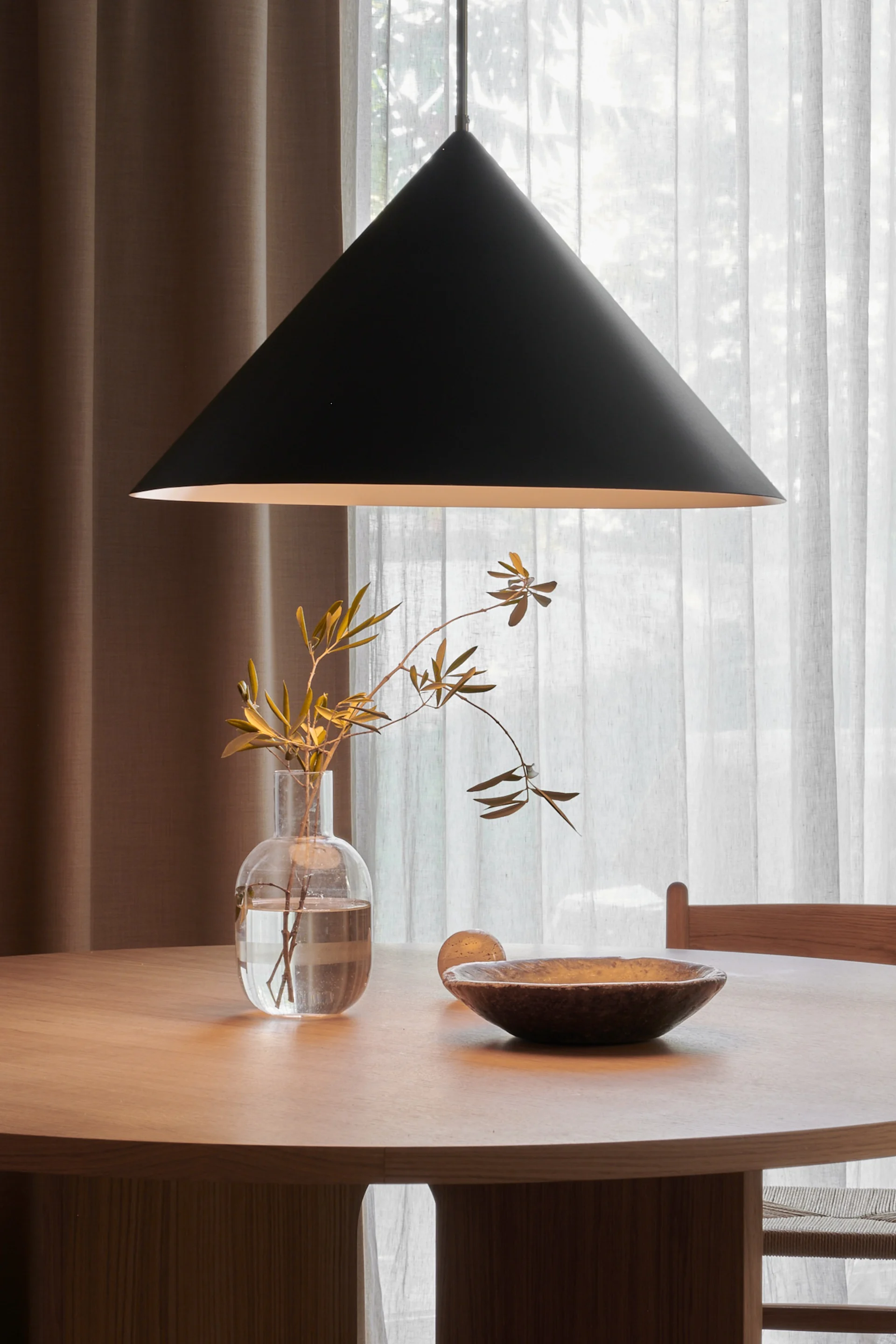 Round wooden dining table with a black conical pendant light overhead, featuring a clear vase with branches and a dark wooden bowl.