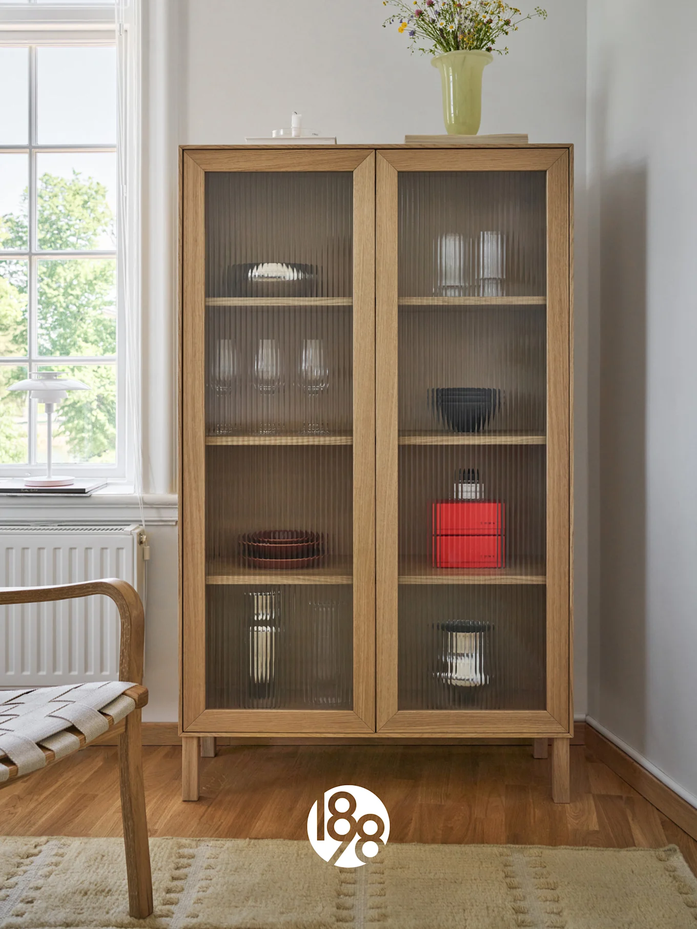 A light wooden cabinet with fluted glass doors displaying dishes and glasses, topped with a vase of wildflowers. A chair and window are to the left.