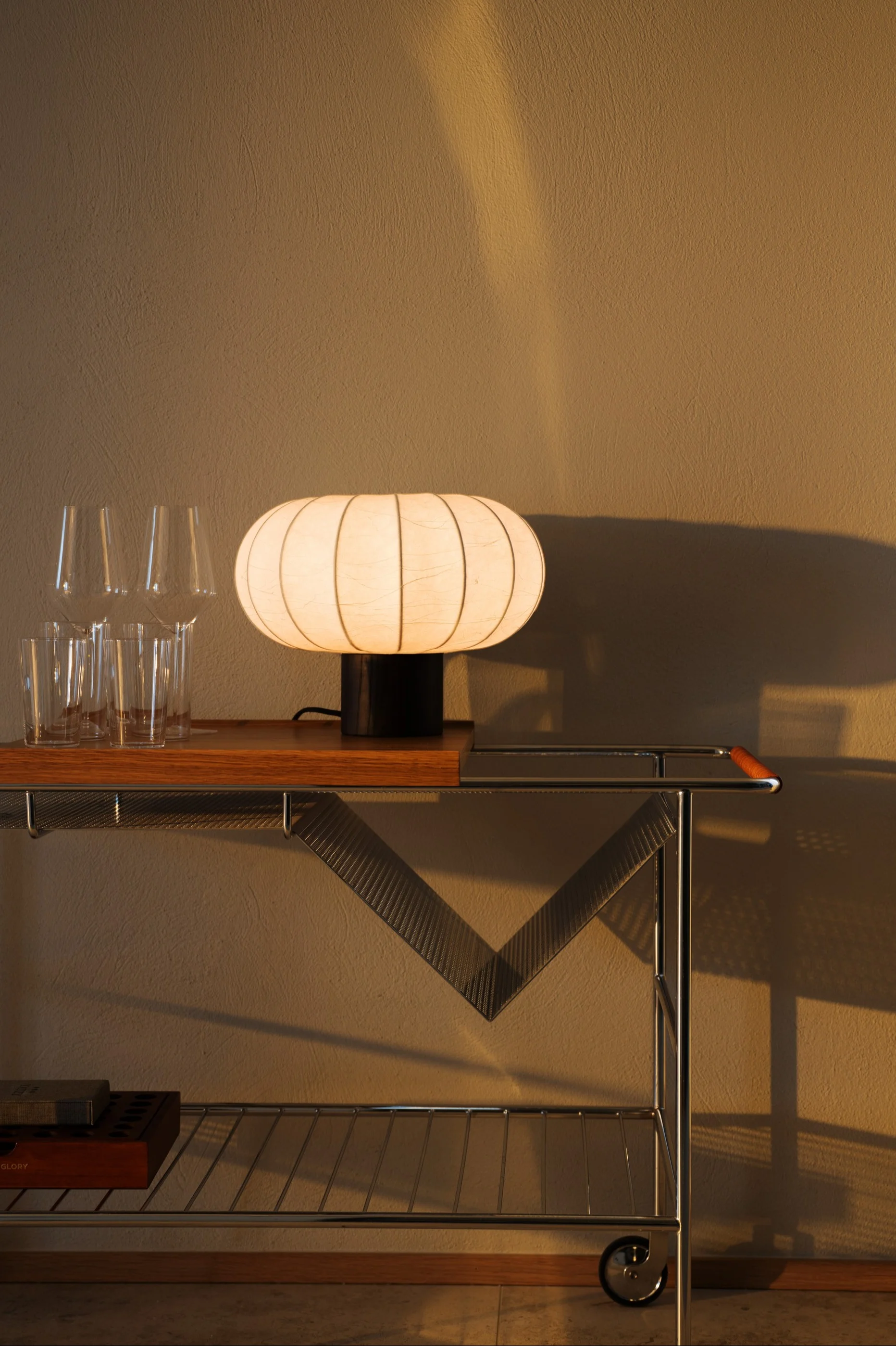 A glowing, ribbed, oval-shaped lamp sits on a wooden and chrome bar cart next to clear glasses, with a textured wall in the background.