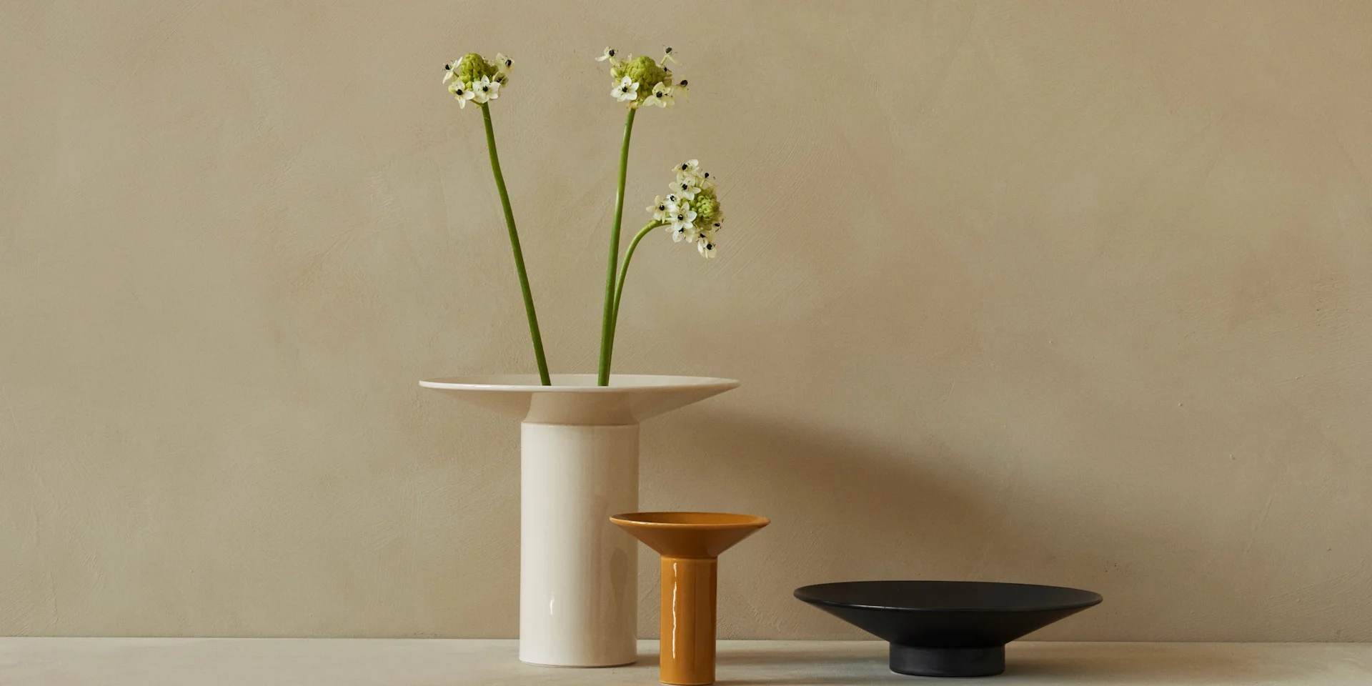 Three ceramic vases in white, amber, and black arranged against a textured beige wall, with white Ornithogalum flowers in the white vase.