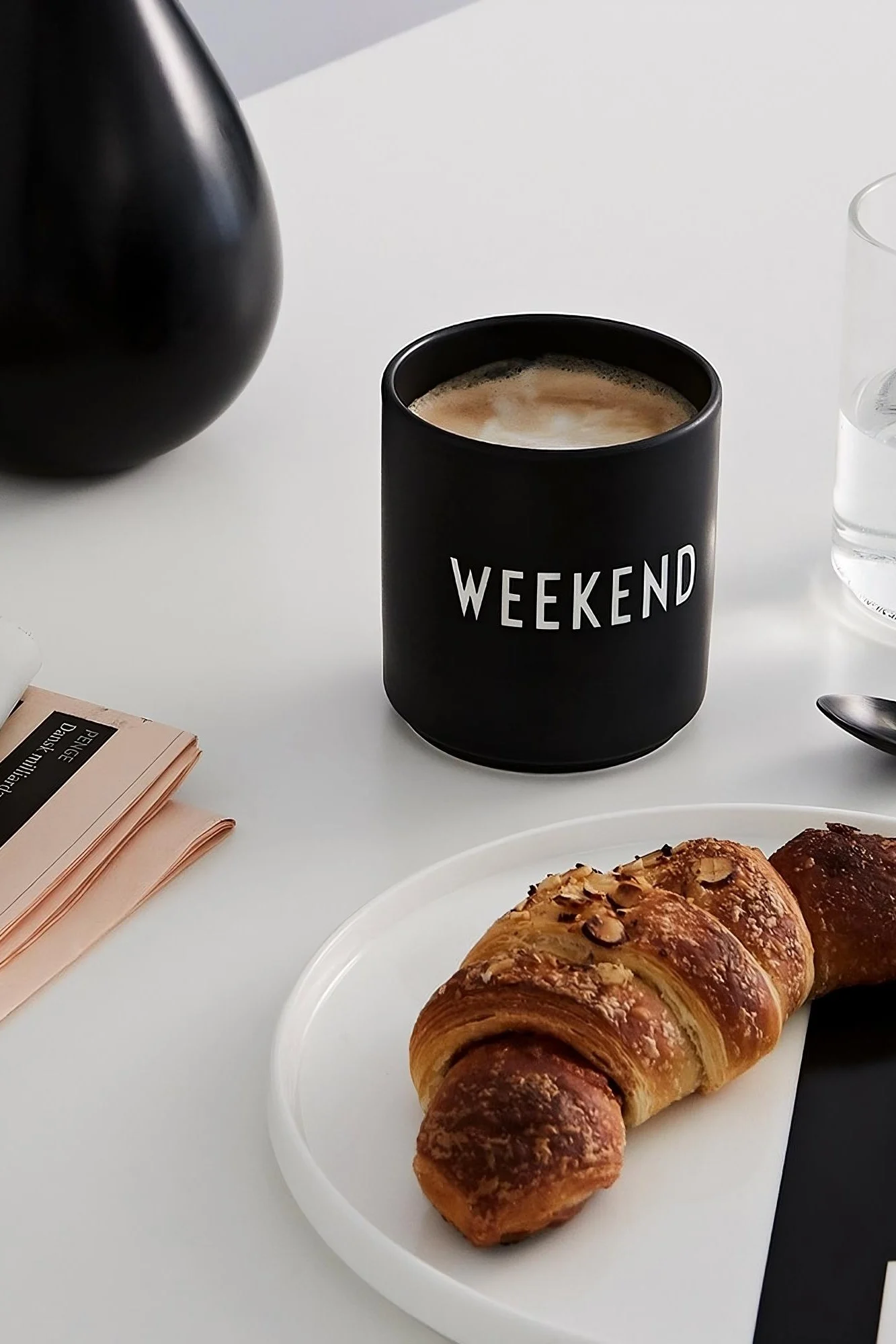Coffee mug: The black coffee mug with the imprint ‘Weekend’ from Design Letters stands on a table, in front of it a plate with a croissant.