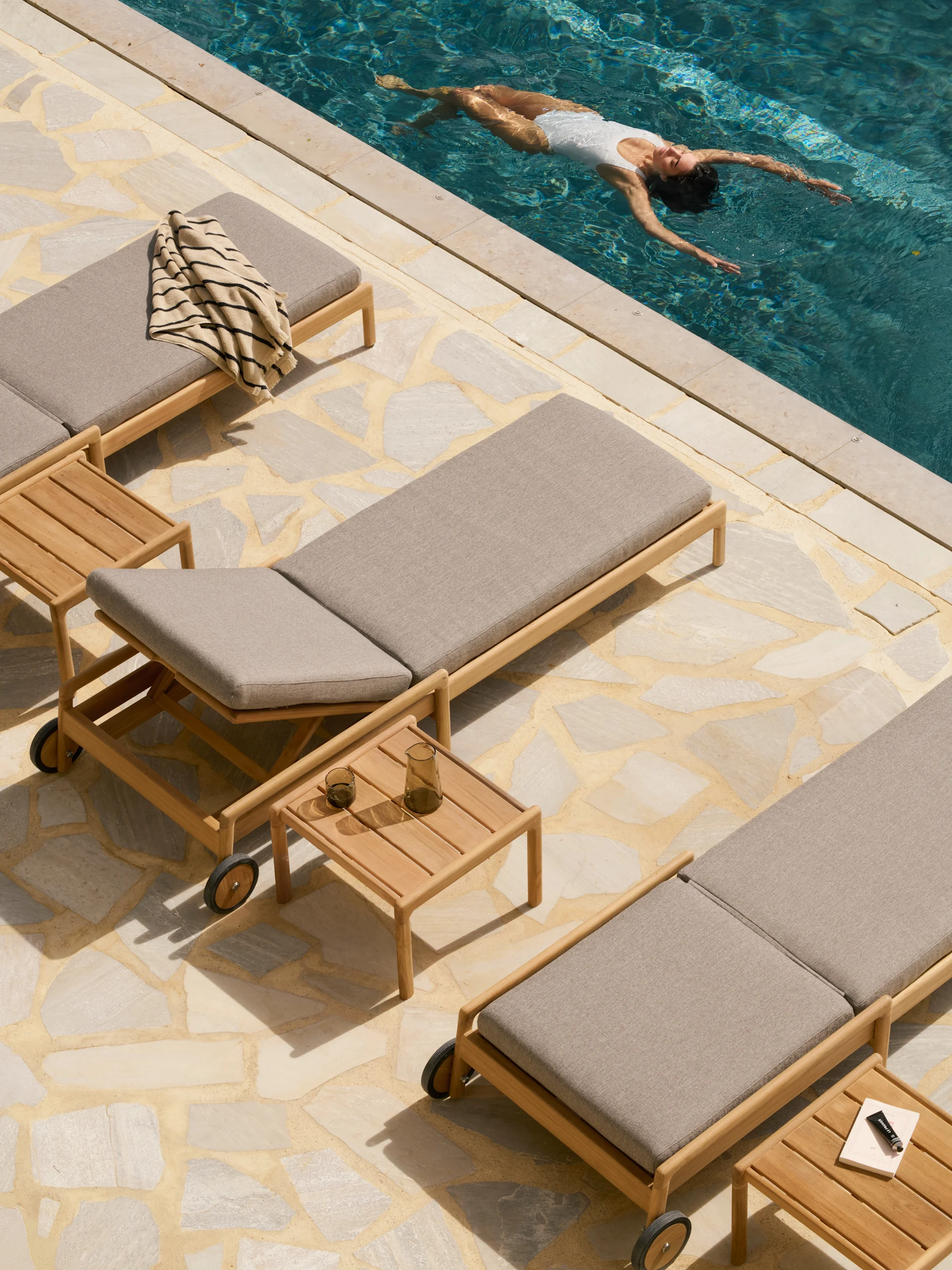 Overhead view of a person floating in a pool surrounded by wooden lounge chairs with grey cushions on a flagstone patio.