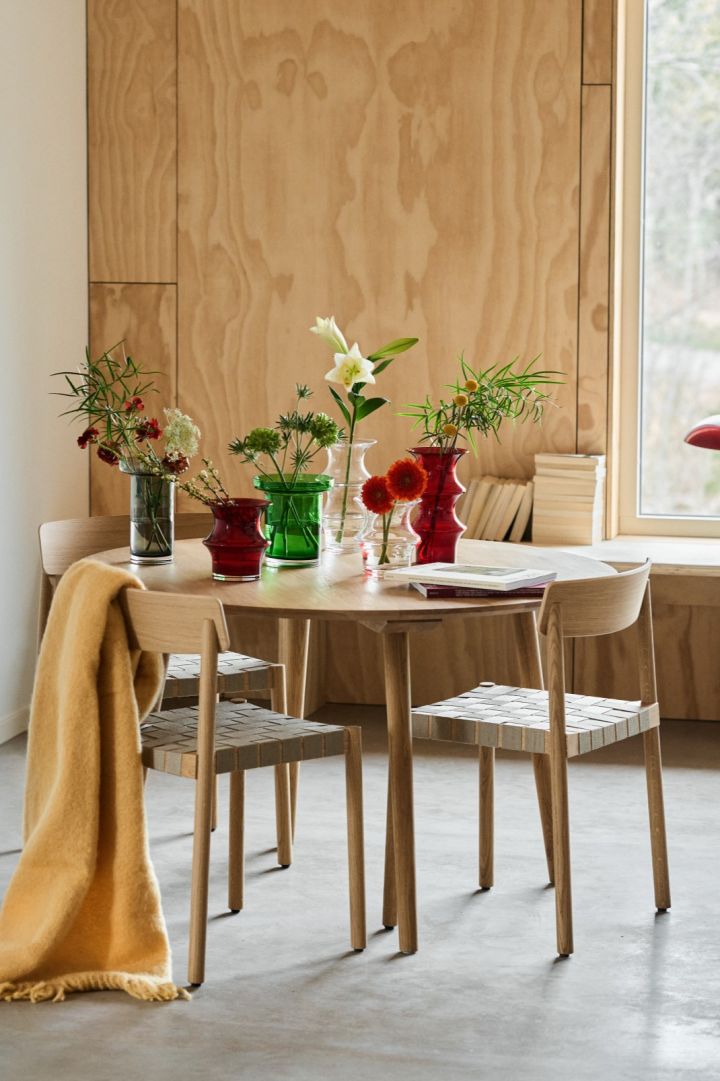 A dining table in the middle of a plywood room with a collection of red, green, grey and clear vases. 