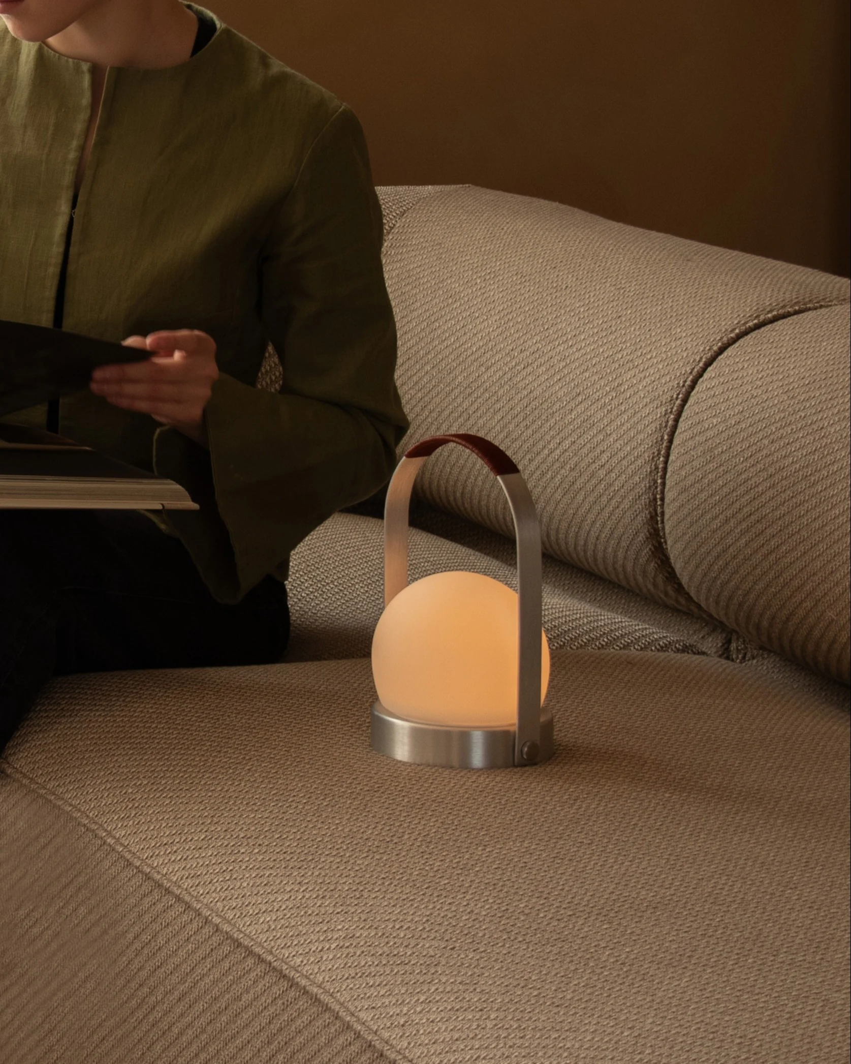A person reads a book on a beige sofa next to a glowing spherical portable lamp with a leather handle.