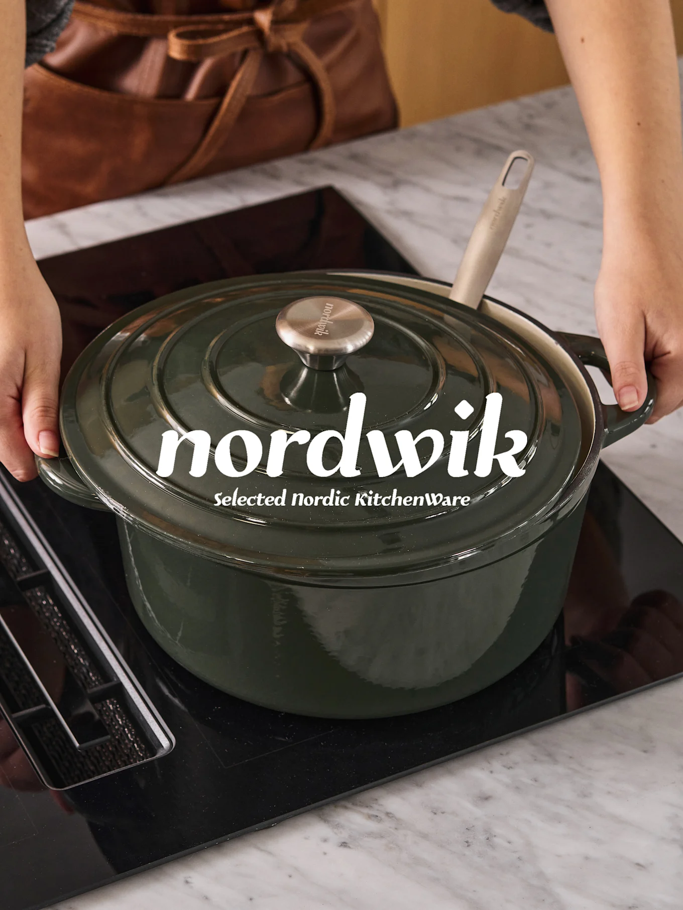 Hands placing a dark green Dutch oven with a light spoon on a black induction cooktop. 'nordwik' is visible on the lid.