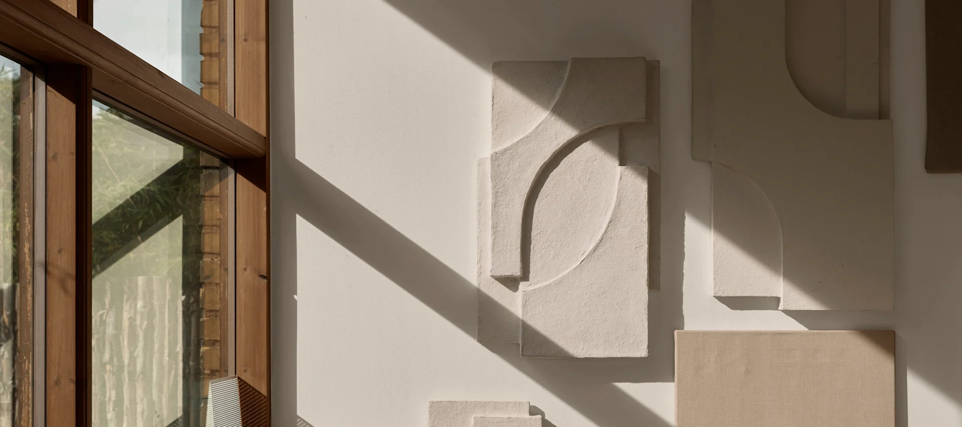 Abstract relief art on a light wall with strong sunlight and diagonal shadows, next to a wooden window frame.