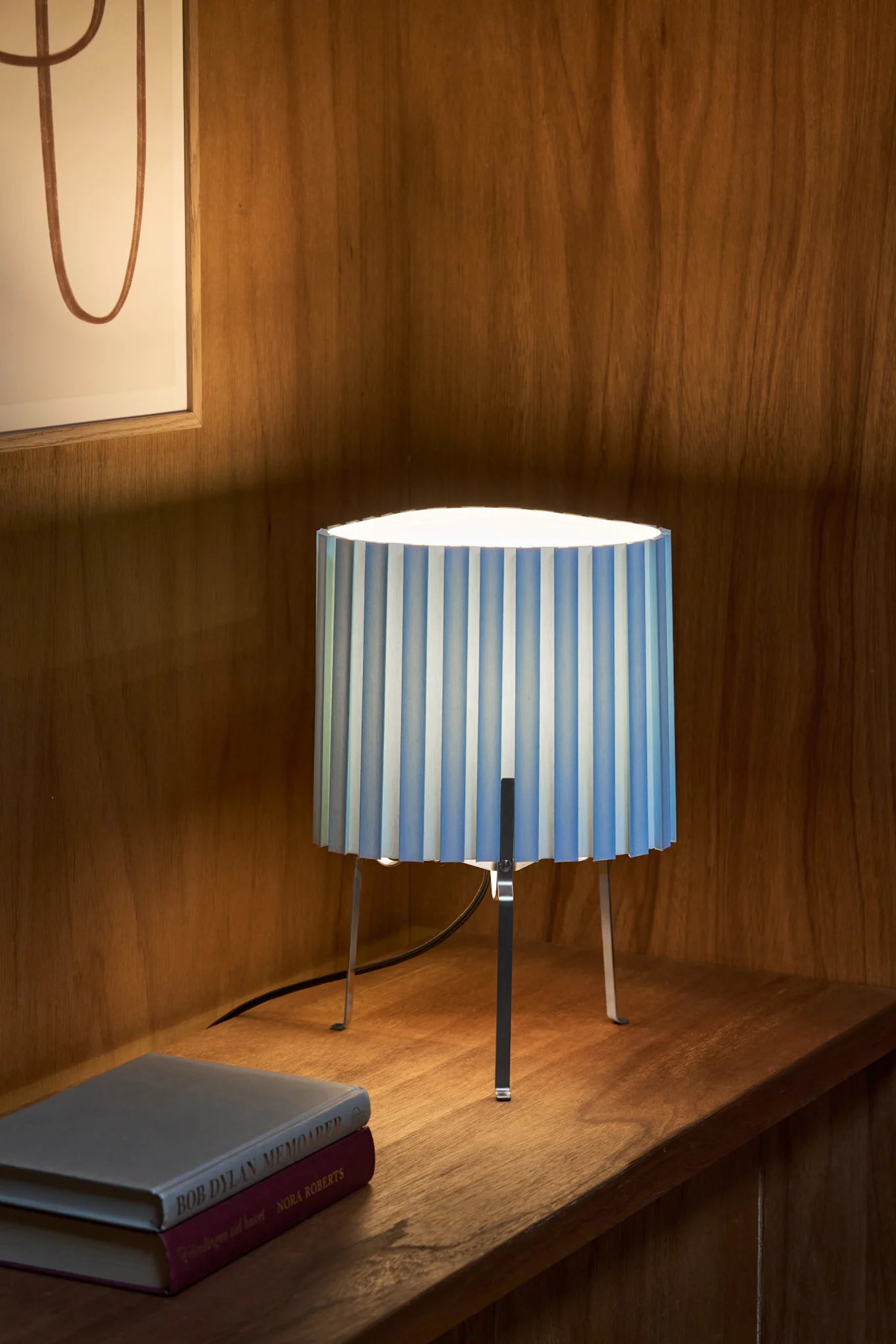 A table lamp with a blue and white striped pleated shade and metal legs, sitting on a wooden shelf next to two books.