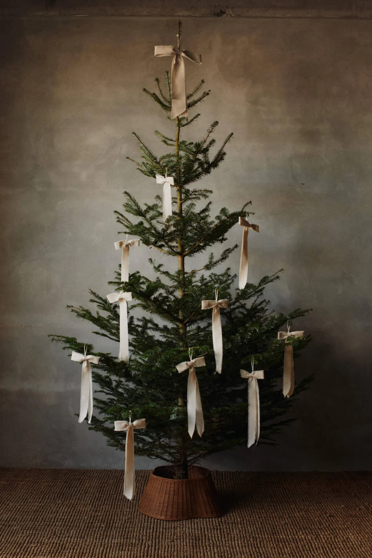 A green Christmas tree in a woven basket, decorated with cream ribbon bows, stands against a textured gray wall on a brown rug.