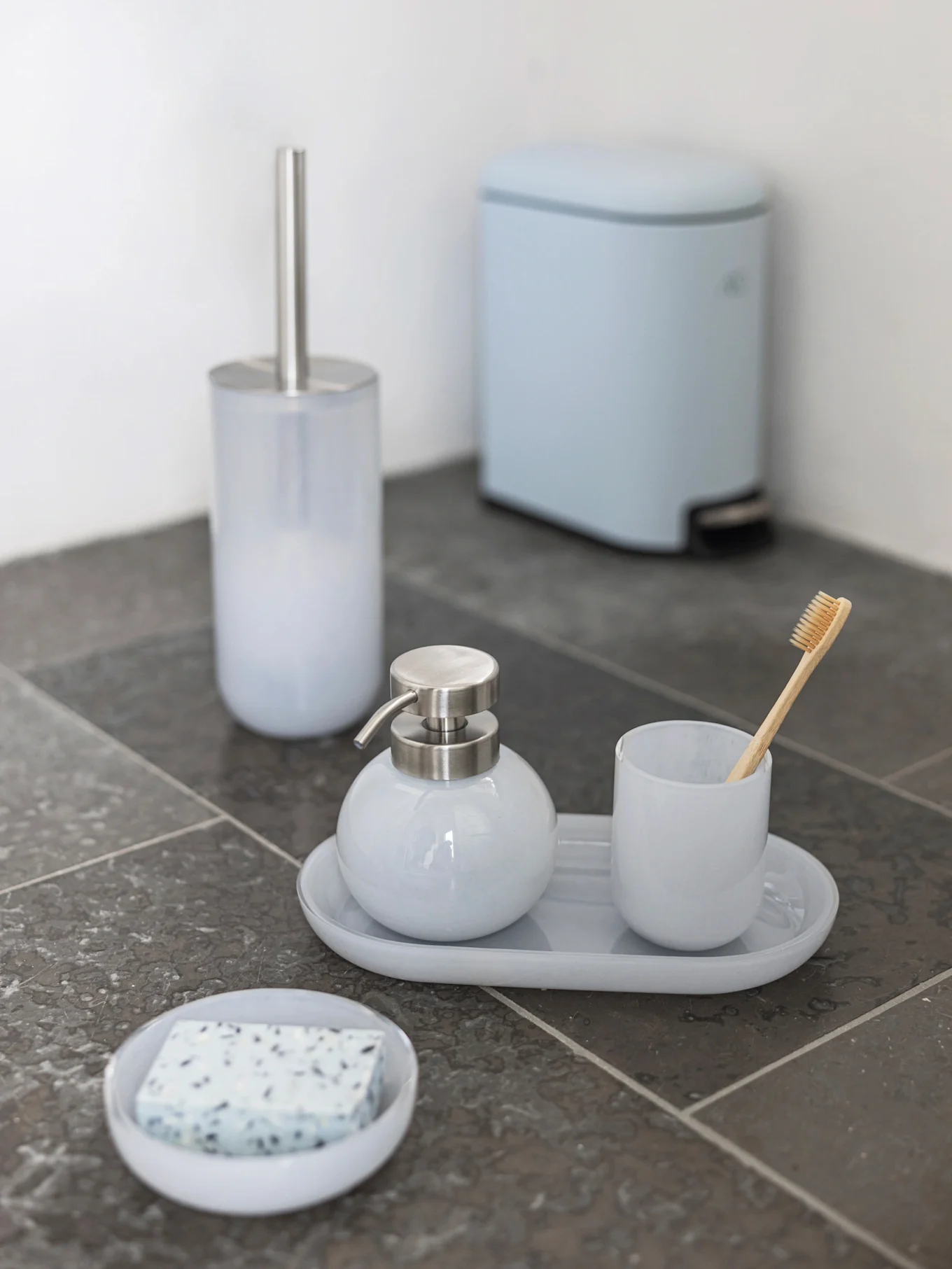 Light blue bathroom set: toilet brush, soap dispenser, cup with bamboo toothbrush, soap dish, and pedal bin on dark tiles.