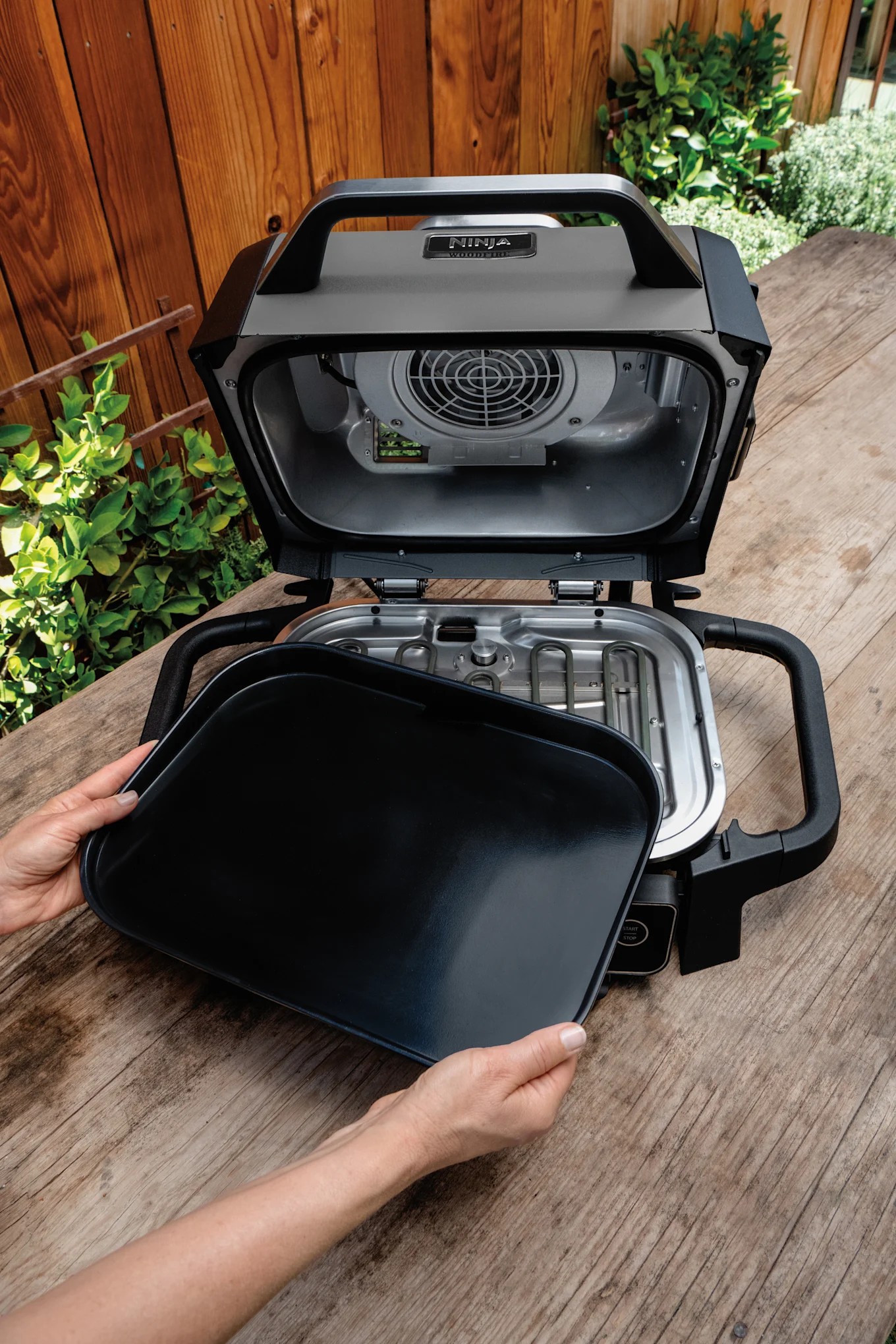 Hands place a griddle into an open black outdoor grill on a wooden table.