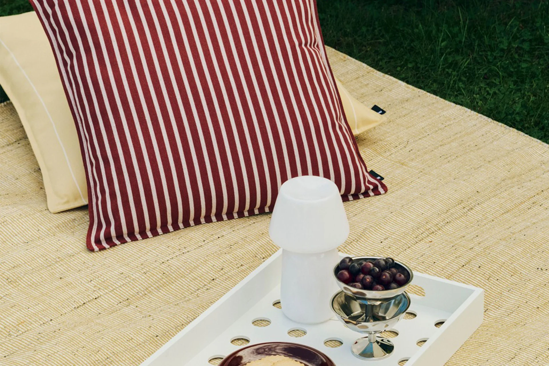Outdoor picnic setup with striped red and white pillow, yellow pillow, white tray holding a mushroom lamp and a bowl of berries.