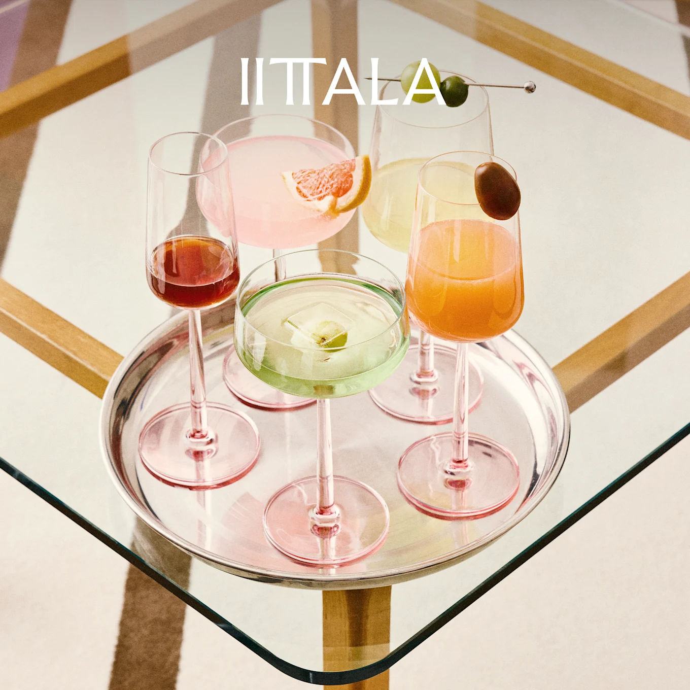 IITTALA logo above a silver tray holding five colorful cocktails in pink-stemmed glasses on a glass table.