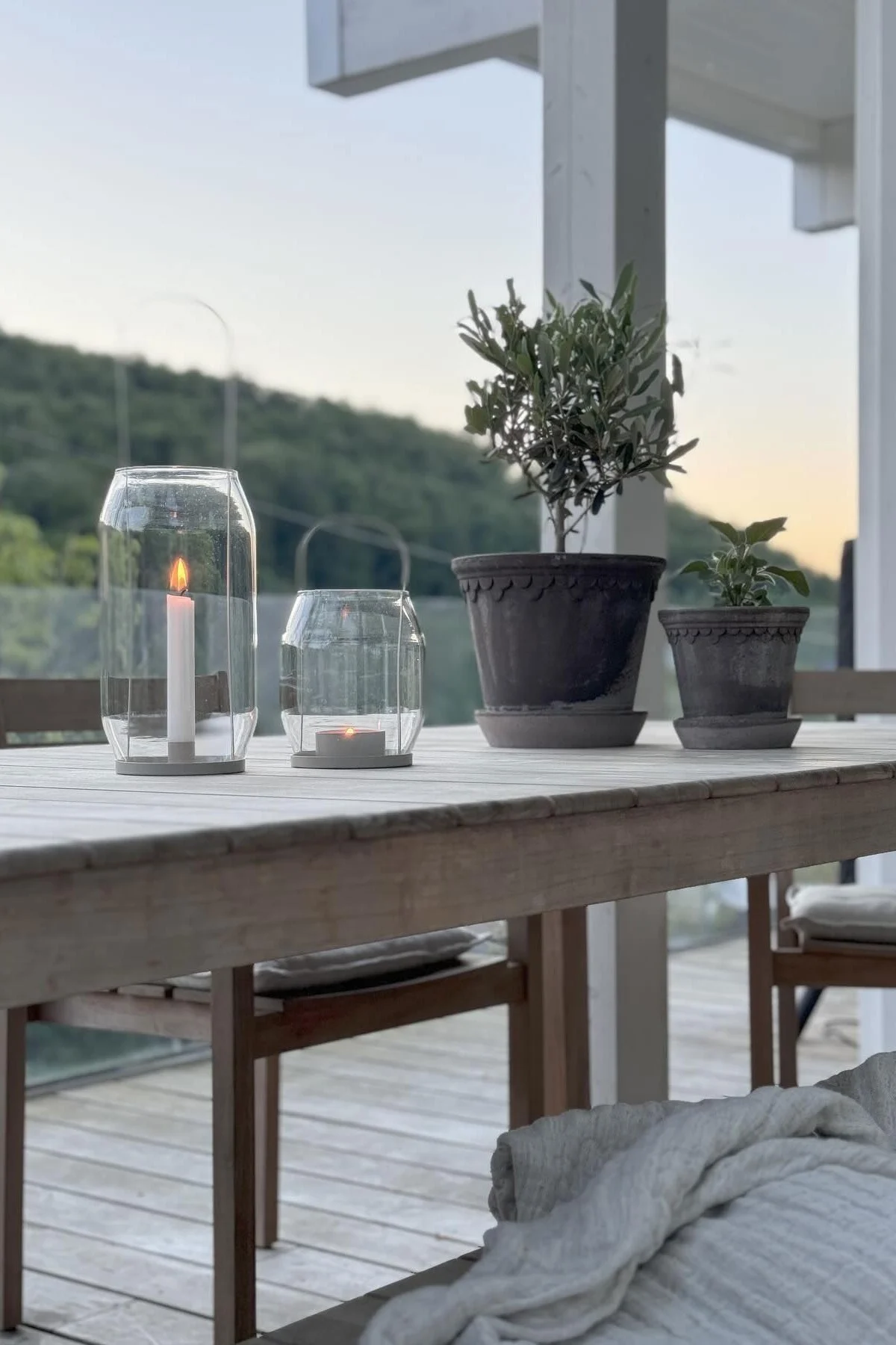 Two lit glass lanterns and two potted plants on a wooden outdoor table at dusk.