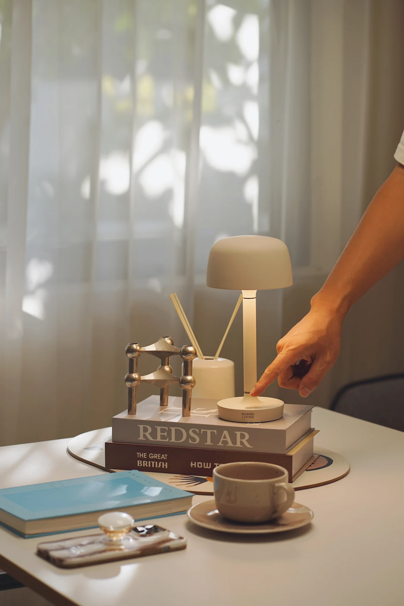 A hand presses a button on a minimalist beige table lamp, casting light on books, a gold candle holder, and a reed diffuser.