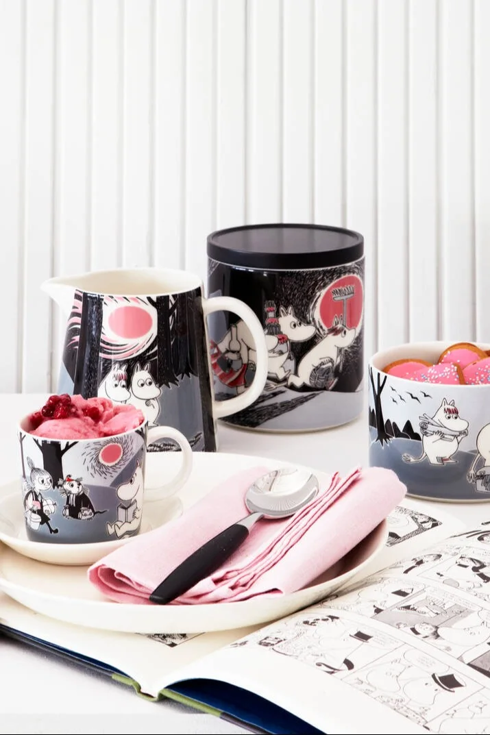 The Moomin Adventure collection, consisting of a cup, plate, jug, tin and bowl, stands on a table.