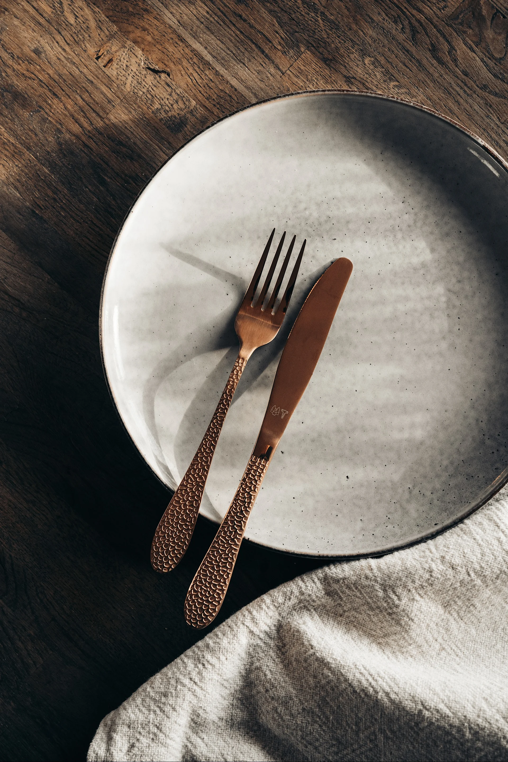 Copper-colored fork and knife with textured handles on a light grey ceramic plate, next to a linen napkin on wood.