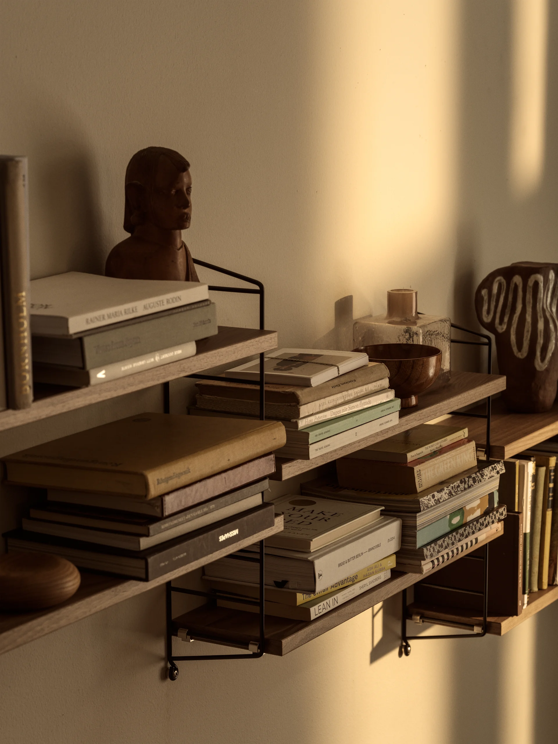 Wall-mounted shelves filled with books, a wooden bust, and decorative objects, illuminated by warm sunlight on the wall.
