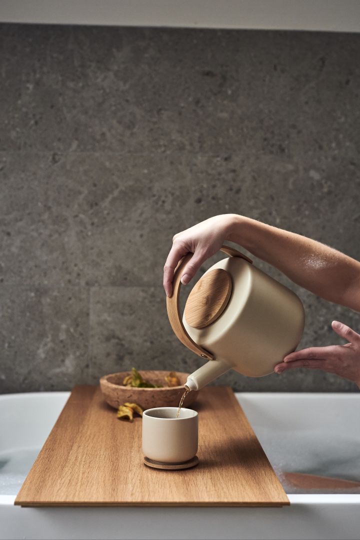 Hands pour tea from a cream white teapot with a wooden lid as part of a home spa session in a warm bubble bath. 