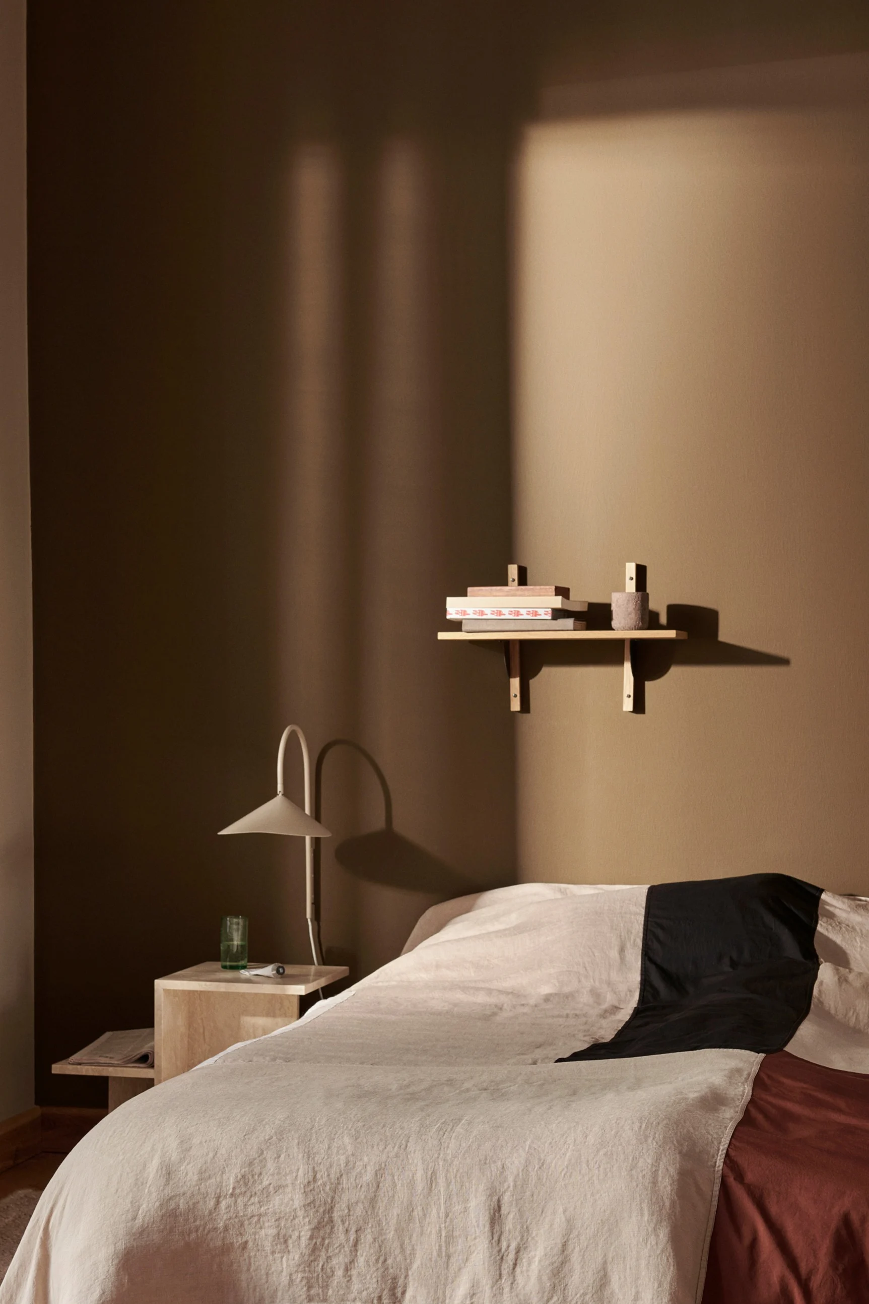A bedroom with a bed featuring beige, black, and rust bedding, a bedside table with a lamp, and a shelf with books on a warm brown wall.