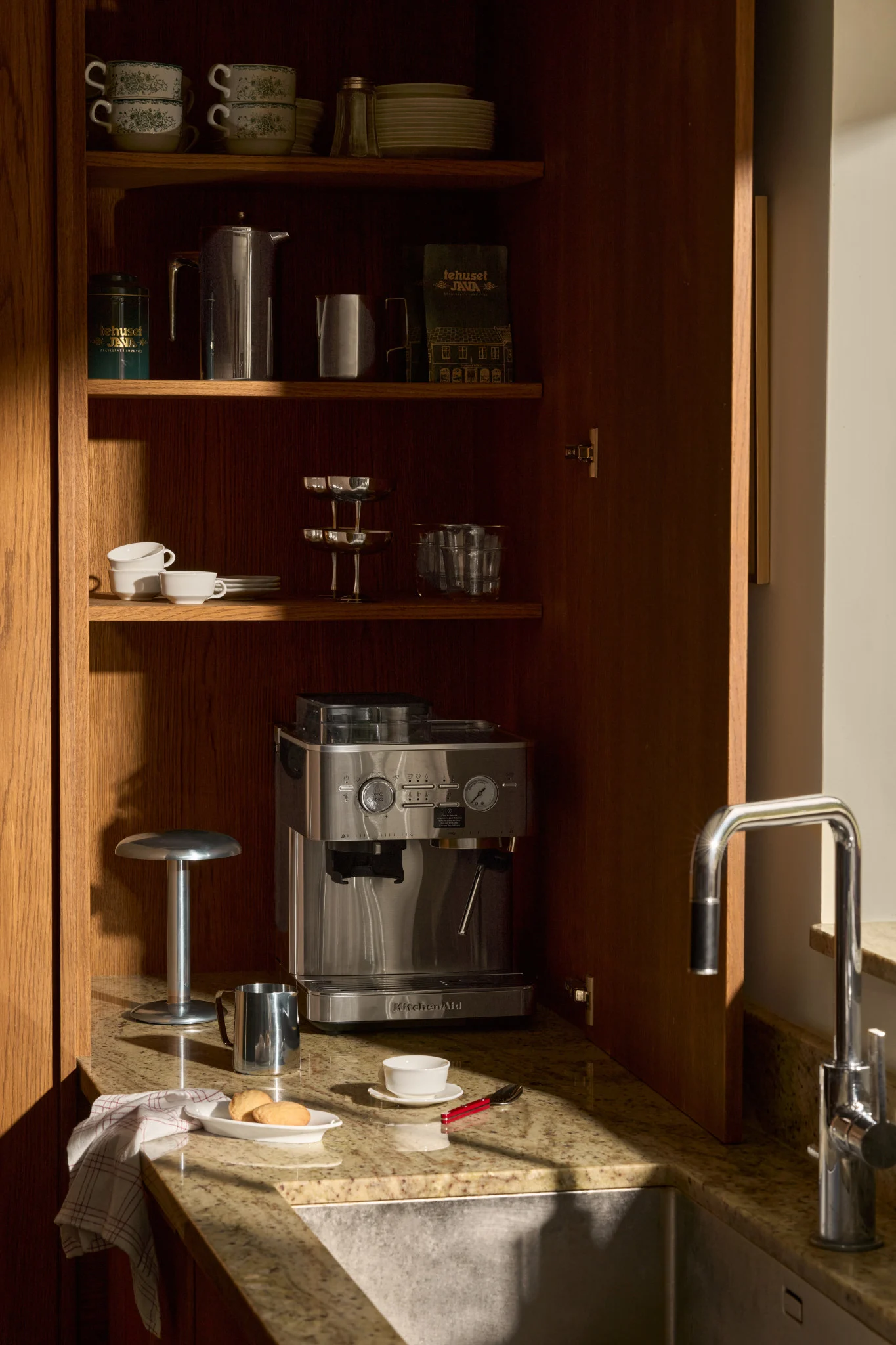 An open wooden kitchen cabinet with an espresso machine, coffee accessories, stacked mugs, and glasses on shelves, next to a sink.