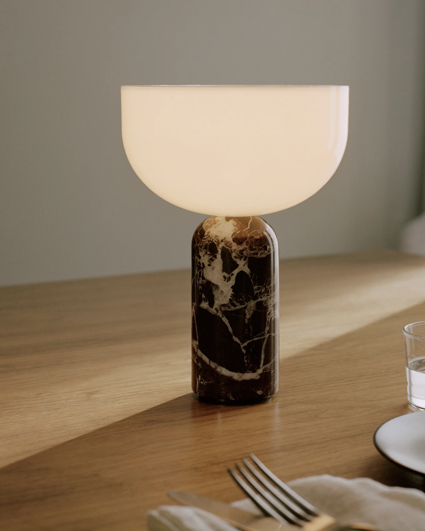 Table lamp with a cylindrical dark marble base with white veins and a semicircular white glass shade that glows, placed on a wooden table with cutlery and a glass.