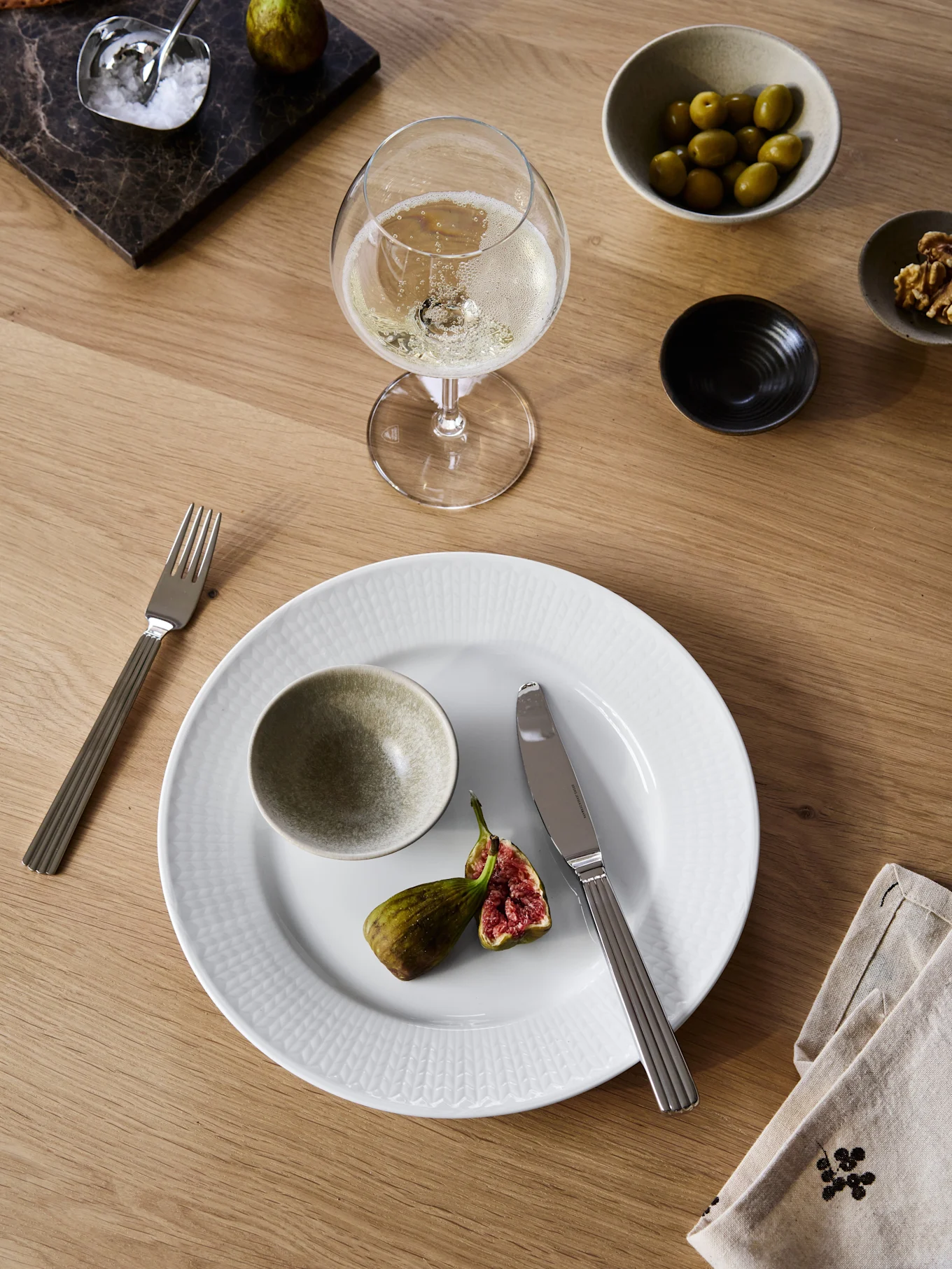Dining table with a white plate, halved fig, fork, knife, sparkling wine, olives, walnuts, crackers, and salt on a marble board.