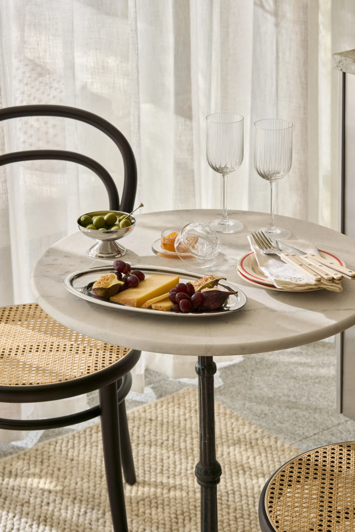 A marble table set with cheese, figs, grapes, olives, jam, glasses, and cutlery, with cane chairs.