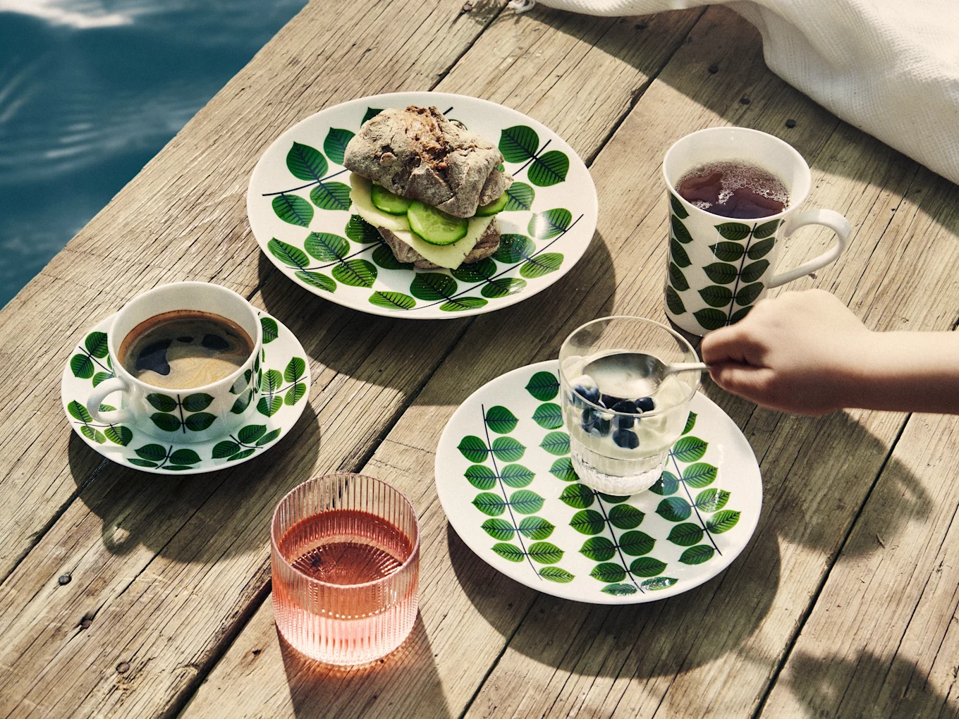 Breakfast on a wooden pier: leaf-patterned plates hold a sandwich, coffee, tea, yogurt with blueberries, and a pink drink.