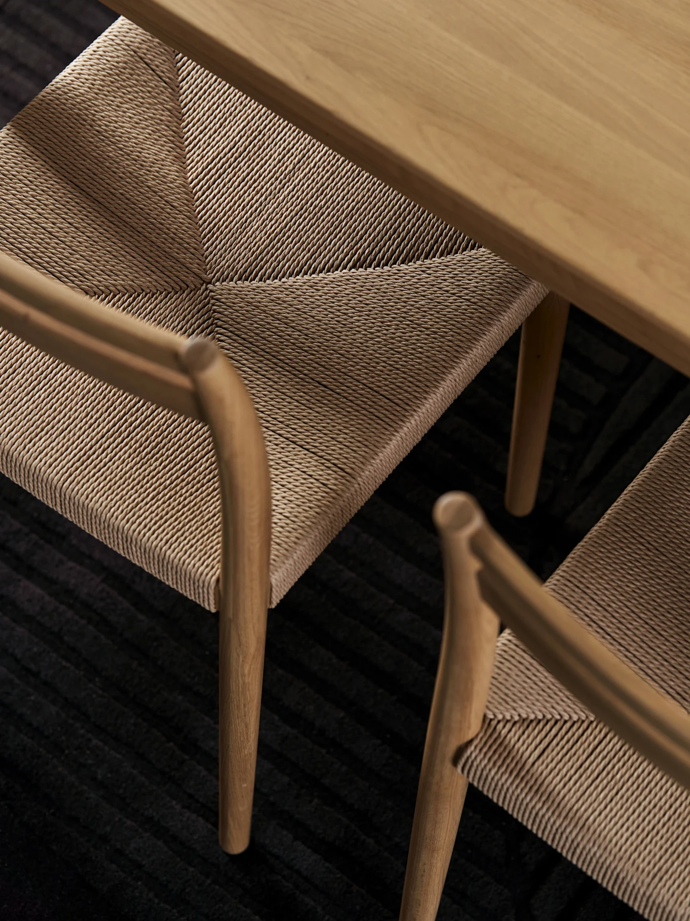 High-angle view of a light wooden table and two chairs with woven seats on a dark ribbed rug.