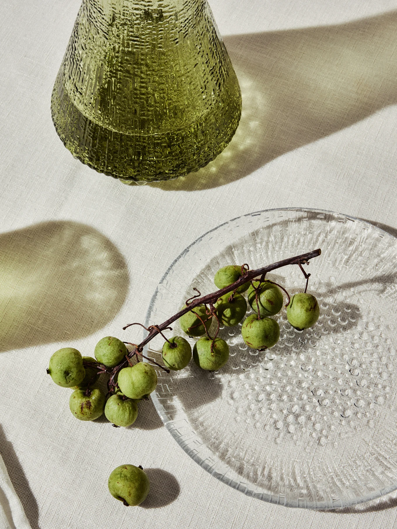 Green textured glass vase and a clear plate with small green fruits on a vine.