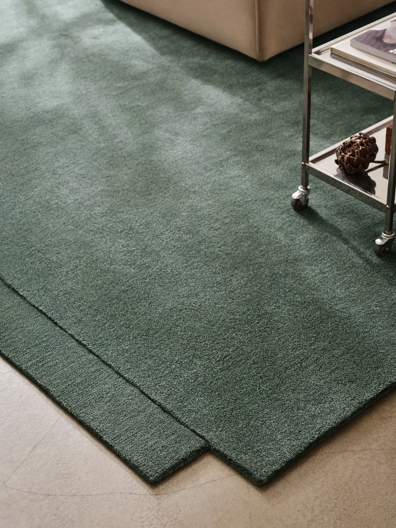 Dark green shag rug on concrete floor, with a beige sofa and metal trolley.