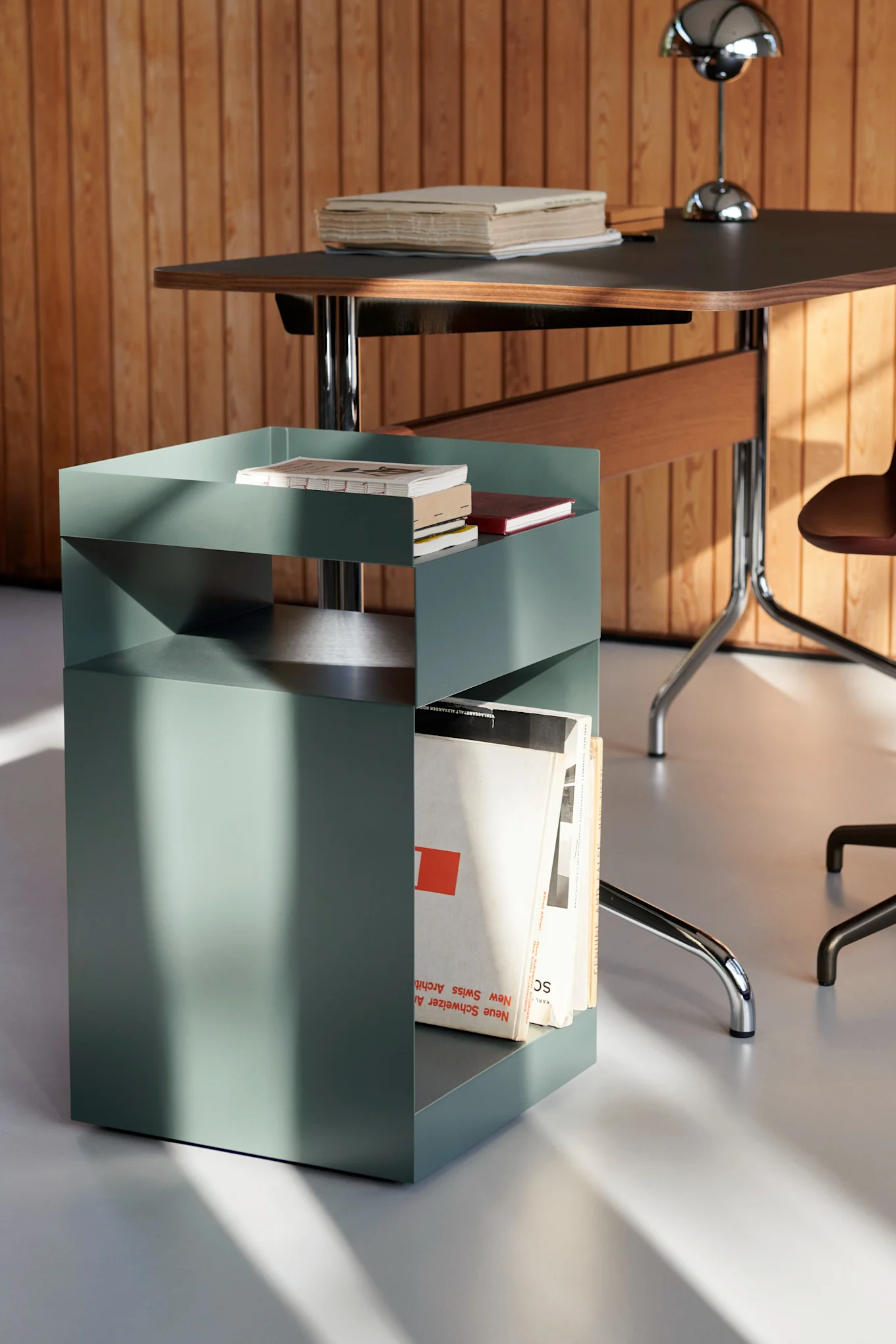Sage green metal side table with geometric design, holding books and magazines, beside a wooden desk with a silver lamp.