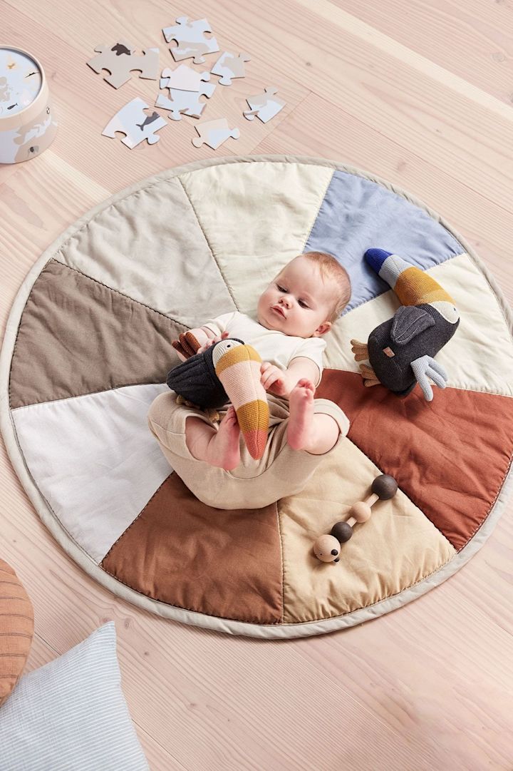 The Trine Tourca cuddly toy from OYOY lies next to a baby on the Moni children's rug. The soft toy is a great and a sustainable baby shower gift.