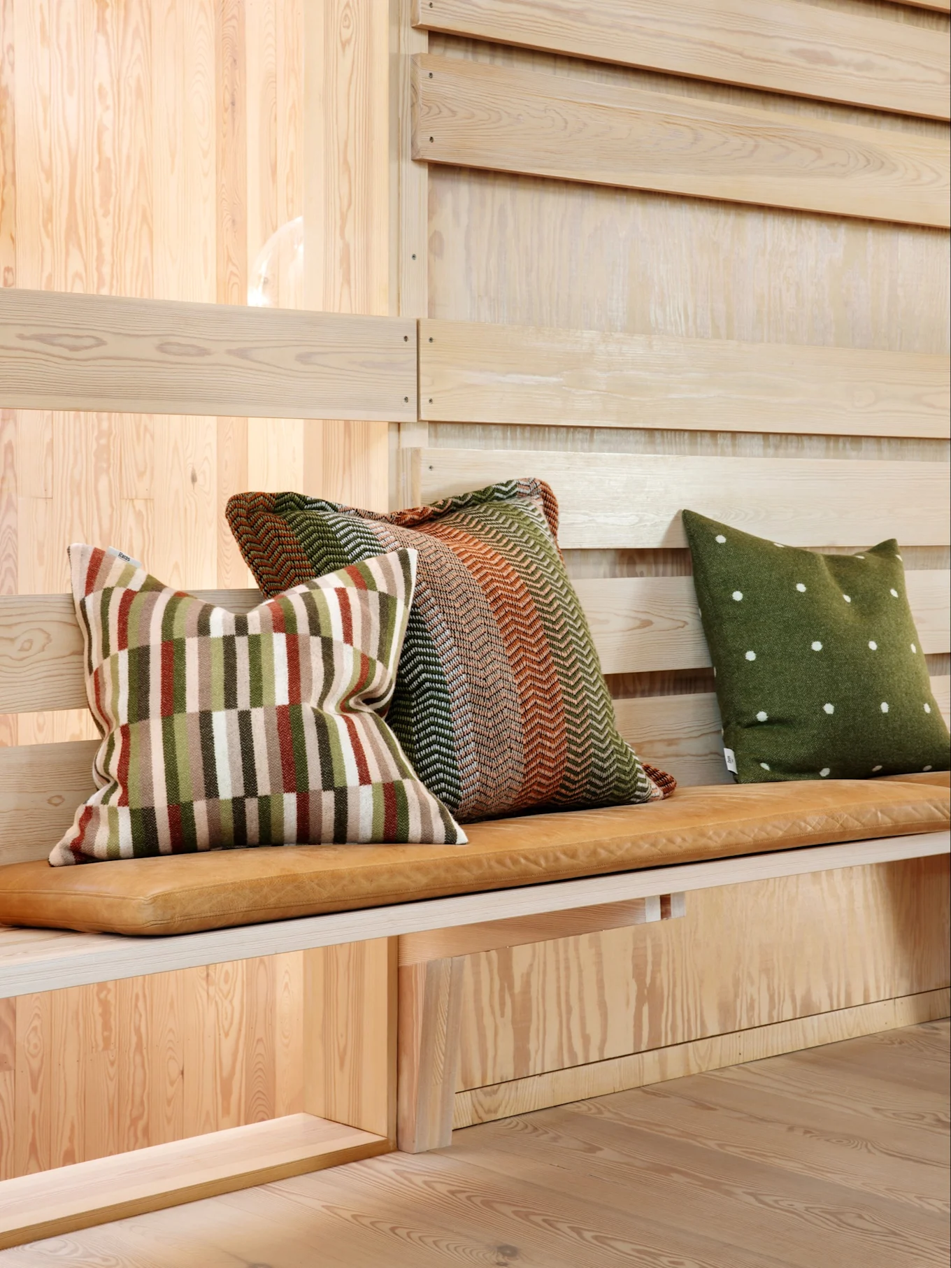 Three decorative pillows on a wooden bench with a leather cushion. One striped, one chevron, and one green with polka dots.