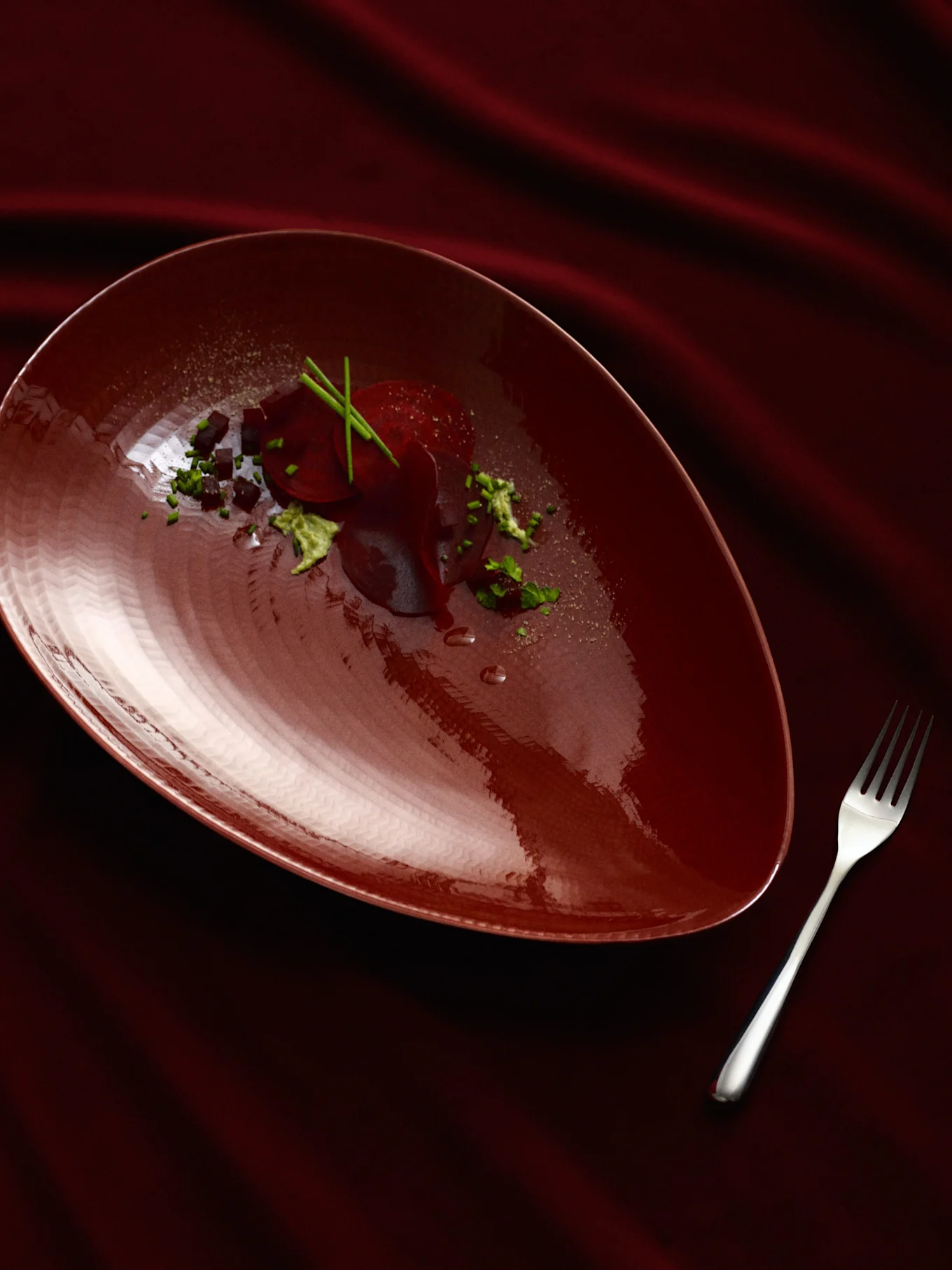 A dark red pear-shaped plate with a beetroot dish, garnished with green herbs and dark cubes, next to a silver fork on a draped dark red fabric.