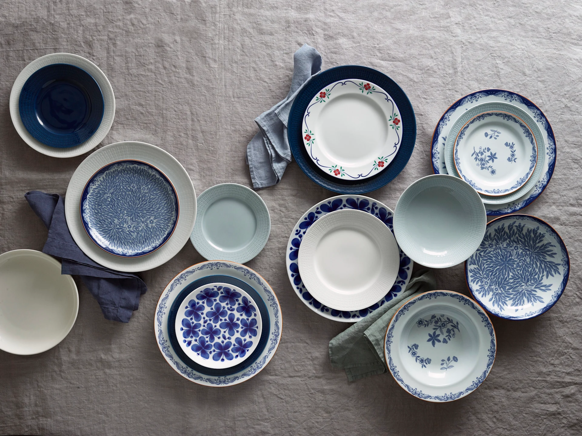 A flat lay of various blue, white, and light grey plates and bowls, some with floral or geometric patterns, arranged with linen napkins on a grey tablecloth.