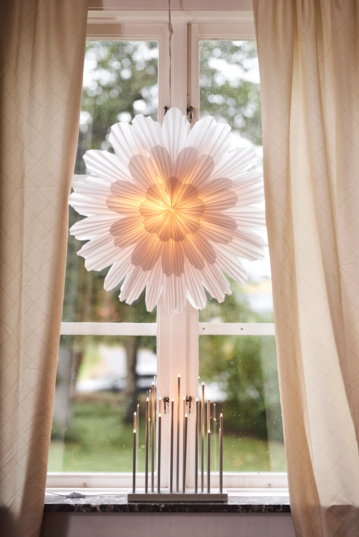 Here you can see a high window framed by curtains, the windowsill is decorated with the Snoblomma Advent star from Watt & Veke and the white Advent holder Elflugan.