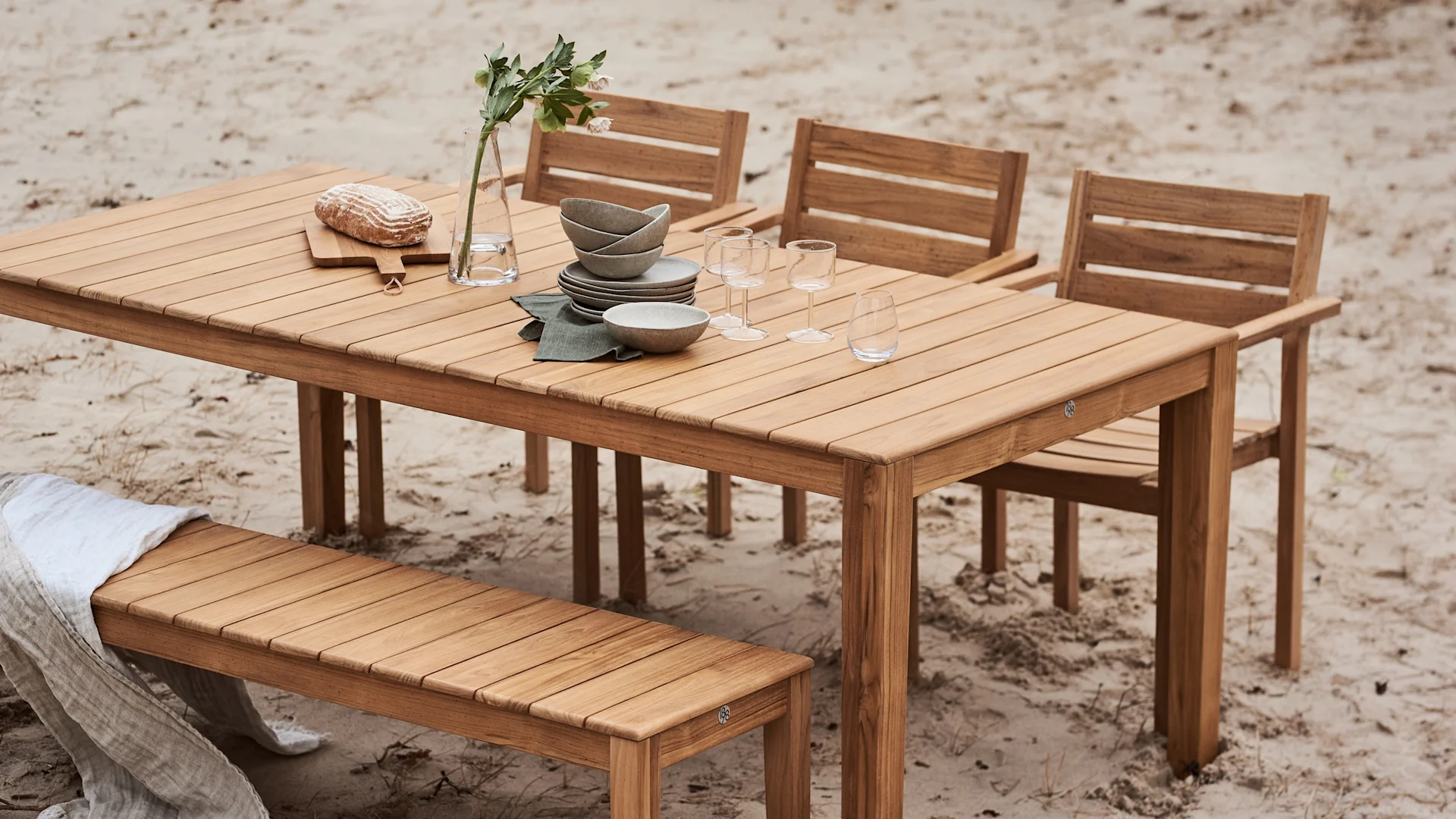 Wooden outdoor dining table set with dishes, glasses, a vase with flowers, and bread, surrounded by chairs and a bench on sand.