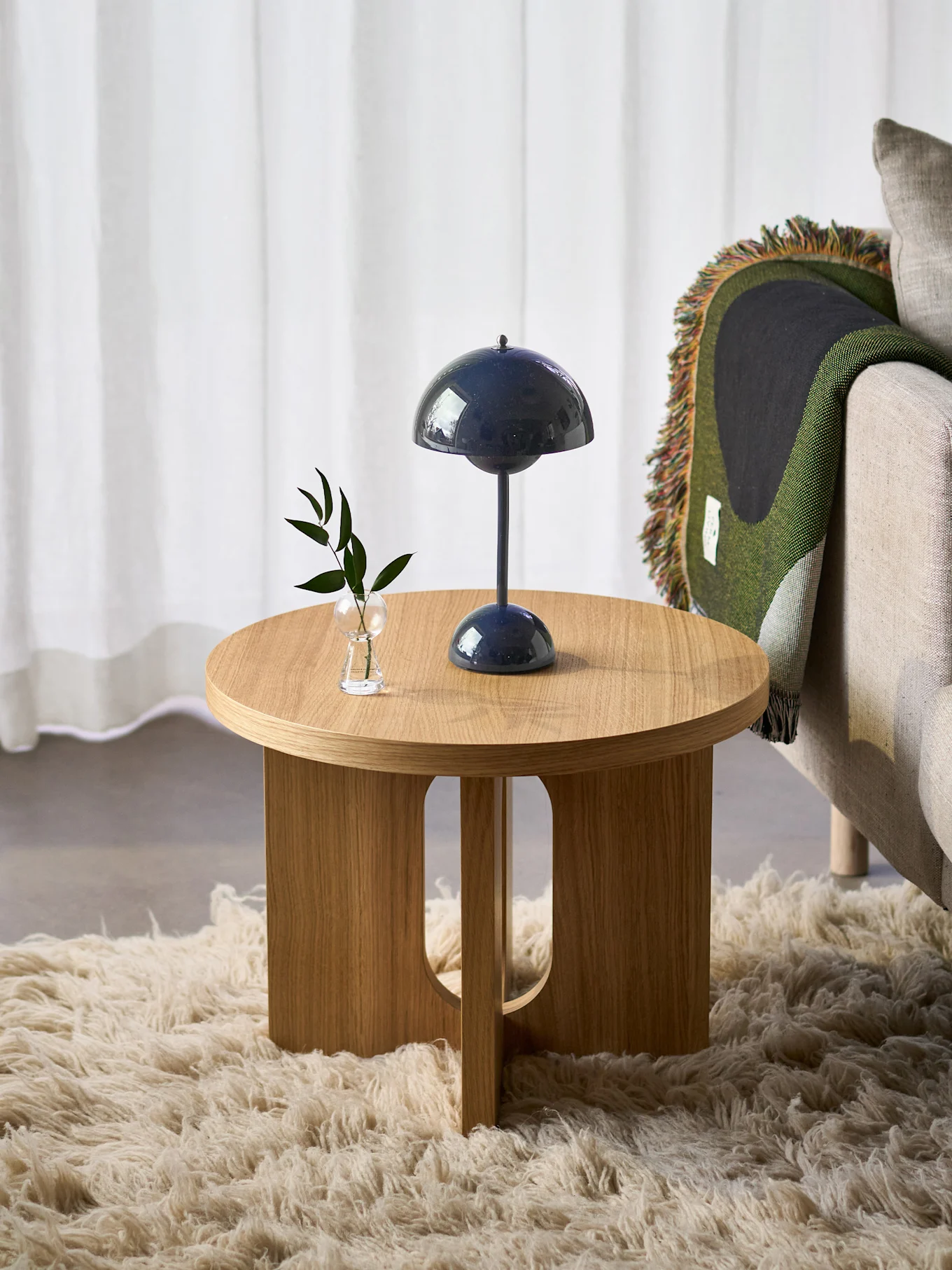A round wooden side table with a dark blue mushroom lamp and a small plant, next to an armchair with a green blanket, on a fluffy rug.